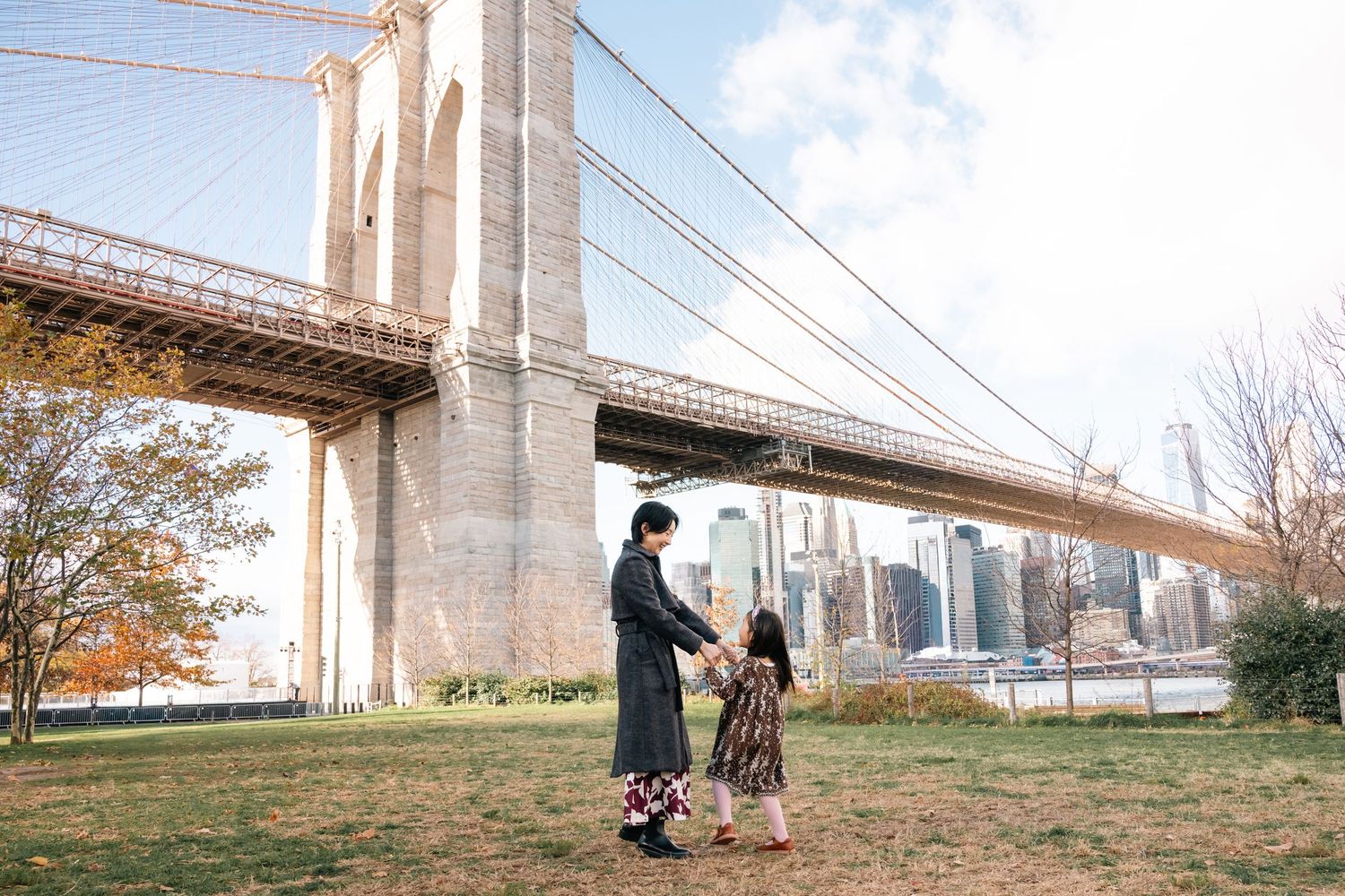 Two people stand together on grass beneath the Brooklyn Bridge with Manhattan skyline in background on autumn day.