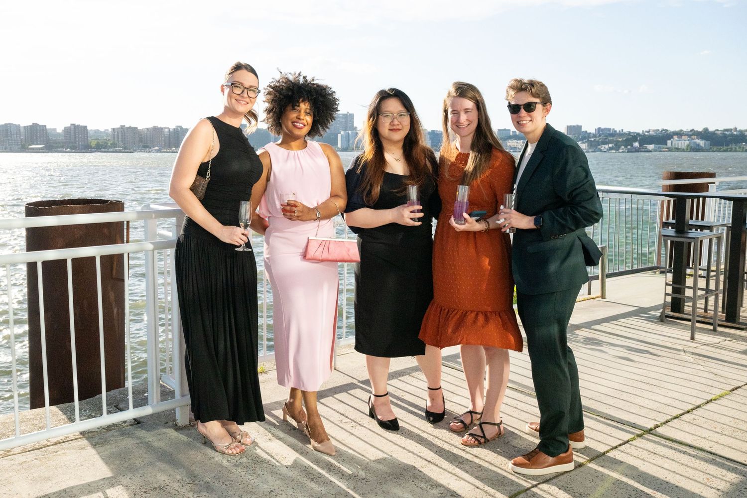 Five women in elegant attire pose together on a waterfront pier during a sunny outdoor event.
