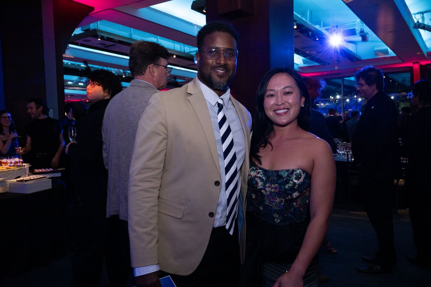 A man in a tan blazer and striped tie poses with a woman in a floral dress at a nighttime event.