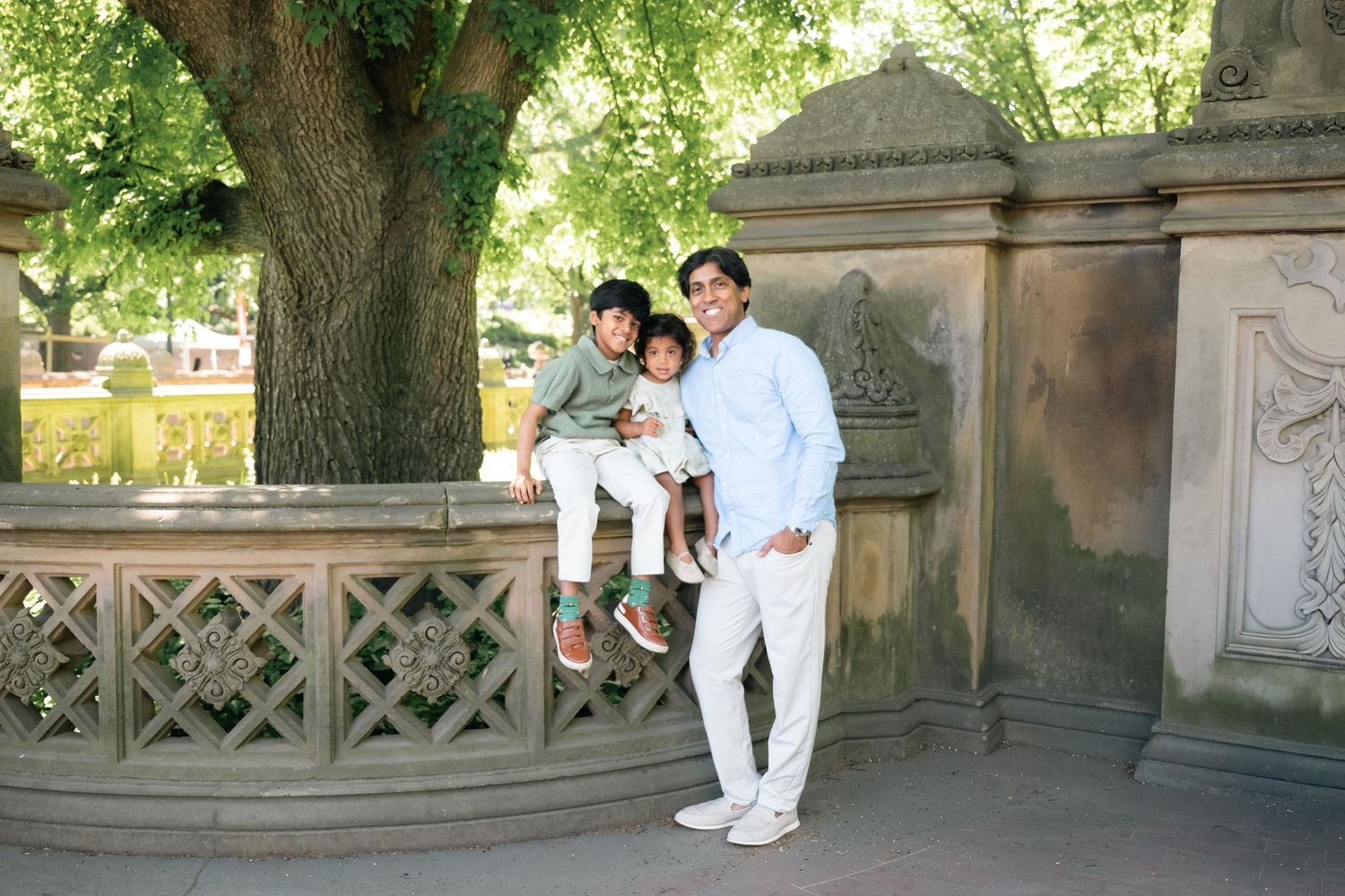 A man poses with two young children on a stone wall in a lush park setting on a sunny day.