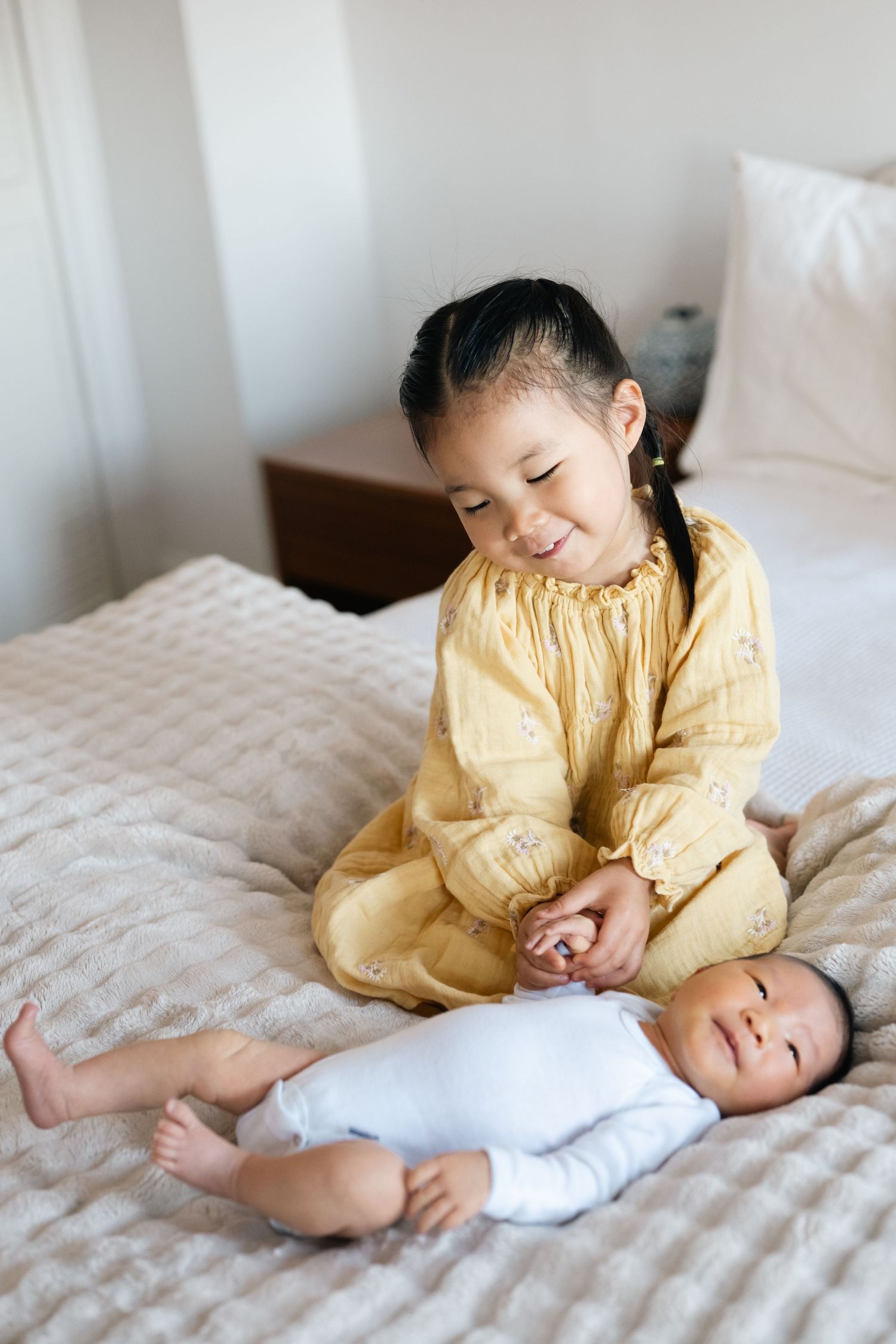 Young child in yellow dress playing sweetly with infant on white bedding.