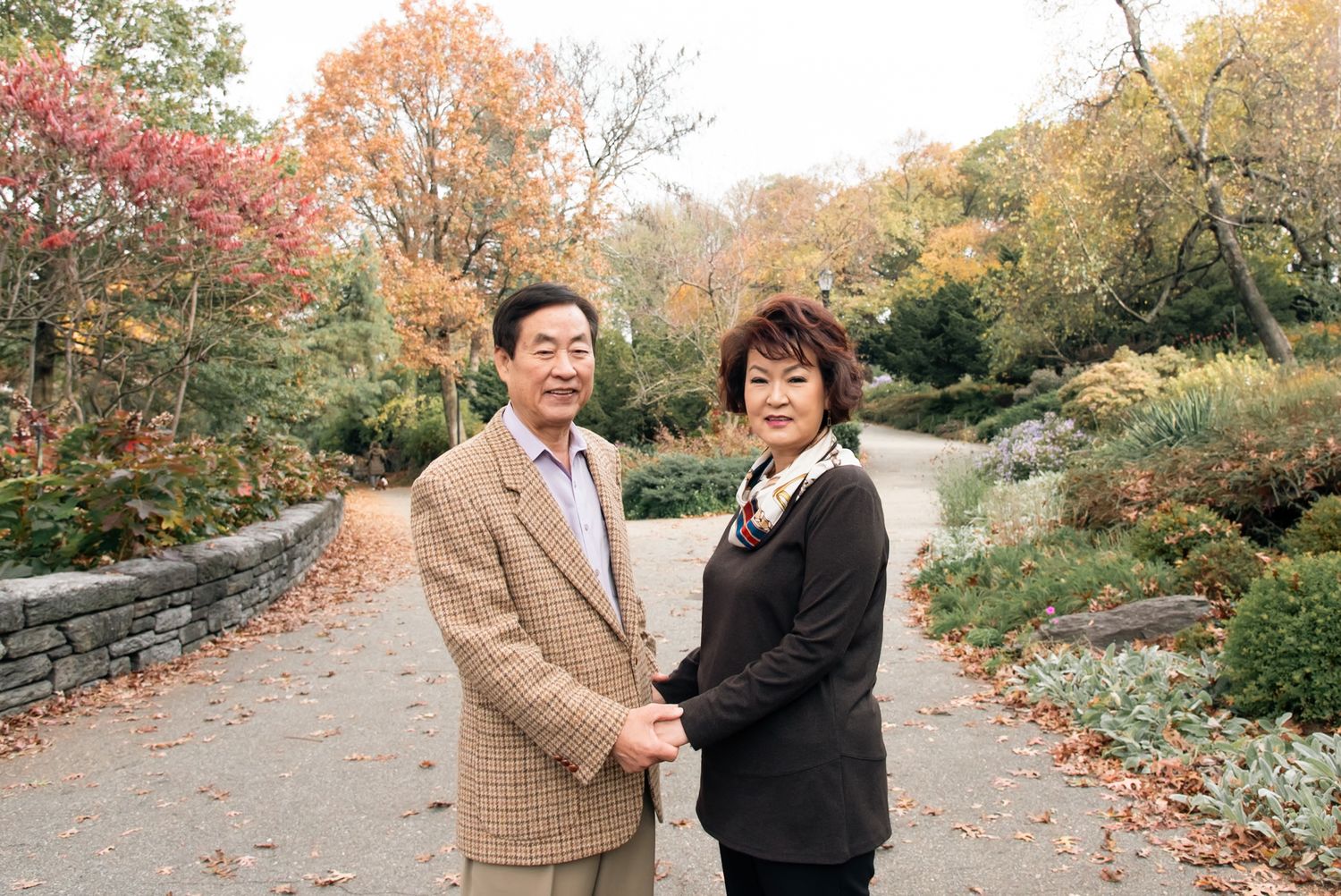 Couple standing together on an autumn path with red maple leaves and stone wall.