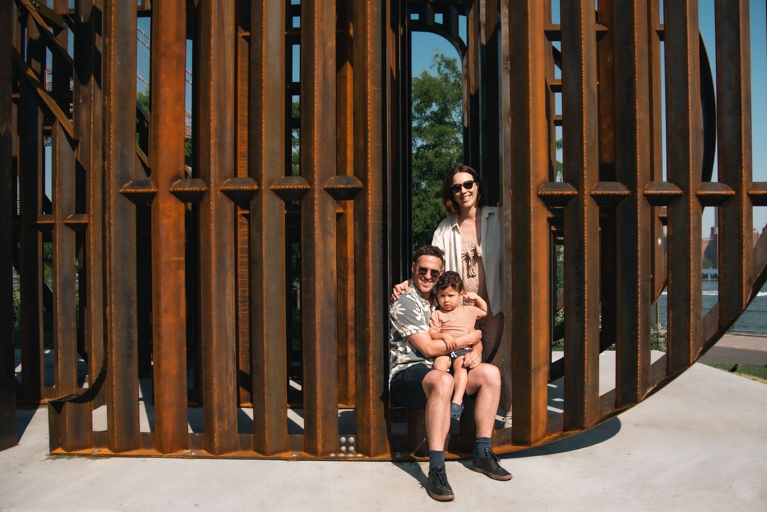 A family of three poses inside a rustic metal sculpture installation outdoors on a sunny day.