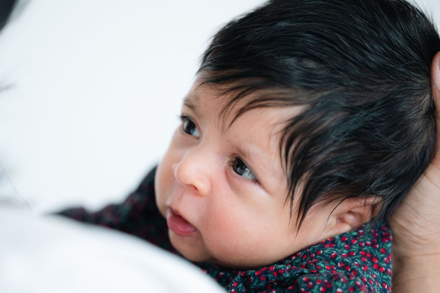 Close-up portrait of a newborn baby with dark hair and alert eyes, resting peacefully in someone's arms.