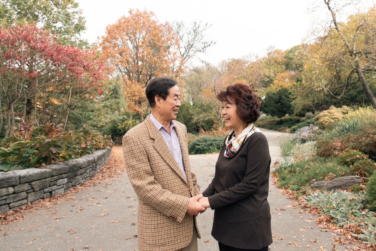 Couple holding hands on a stone-lined path with fall foliage in the background.