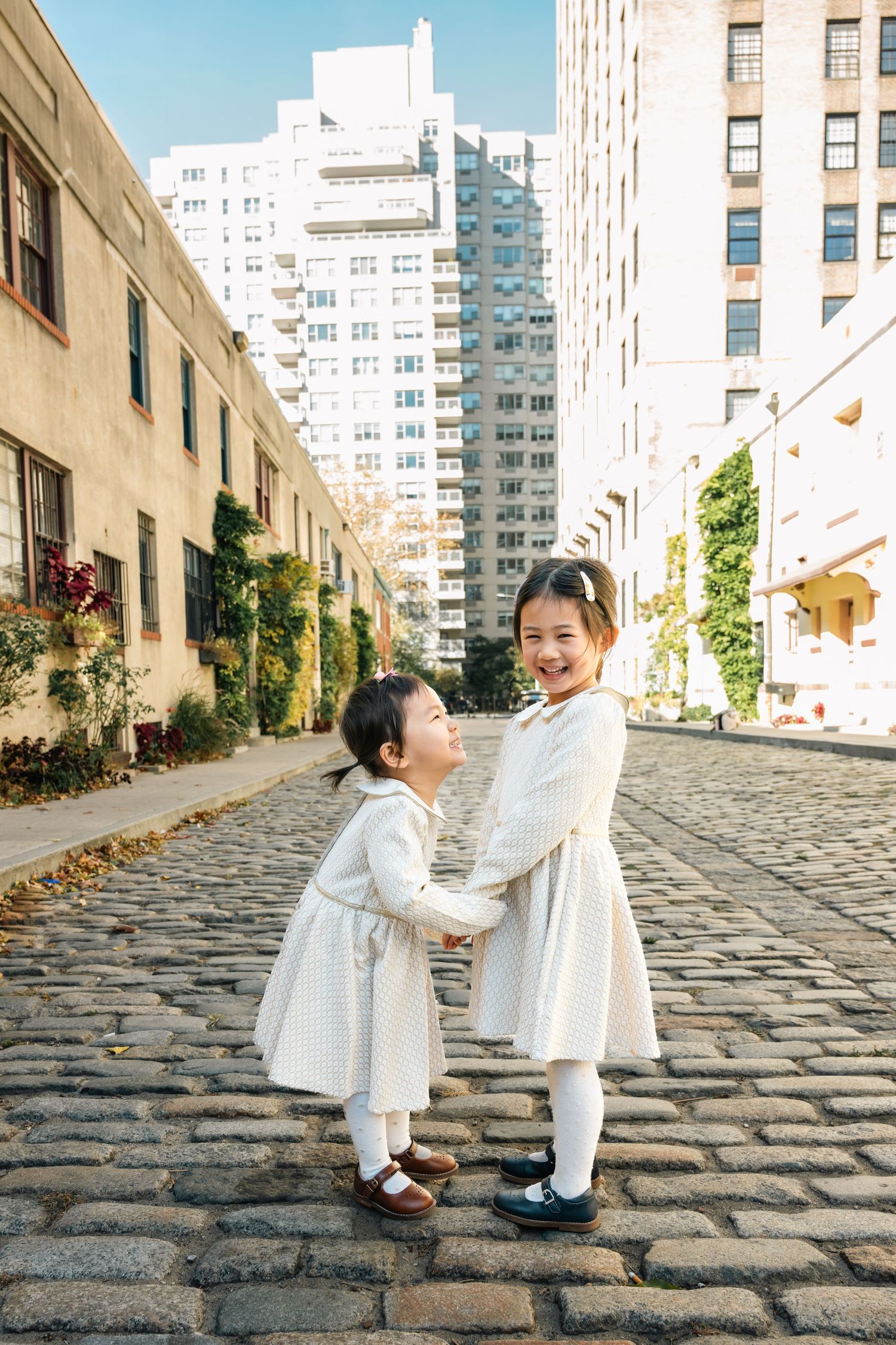 Two small figures in white dresses play together on a cobblestone street with city buildings in the background.