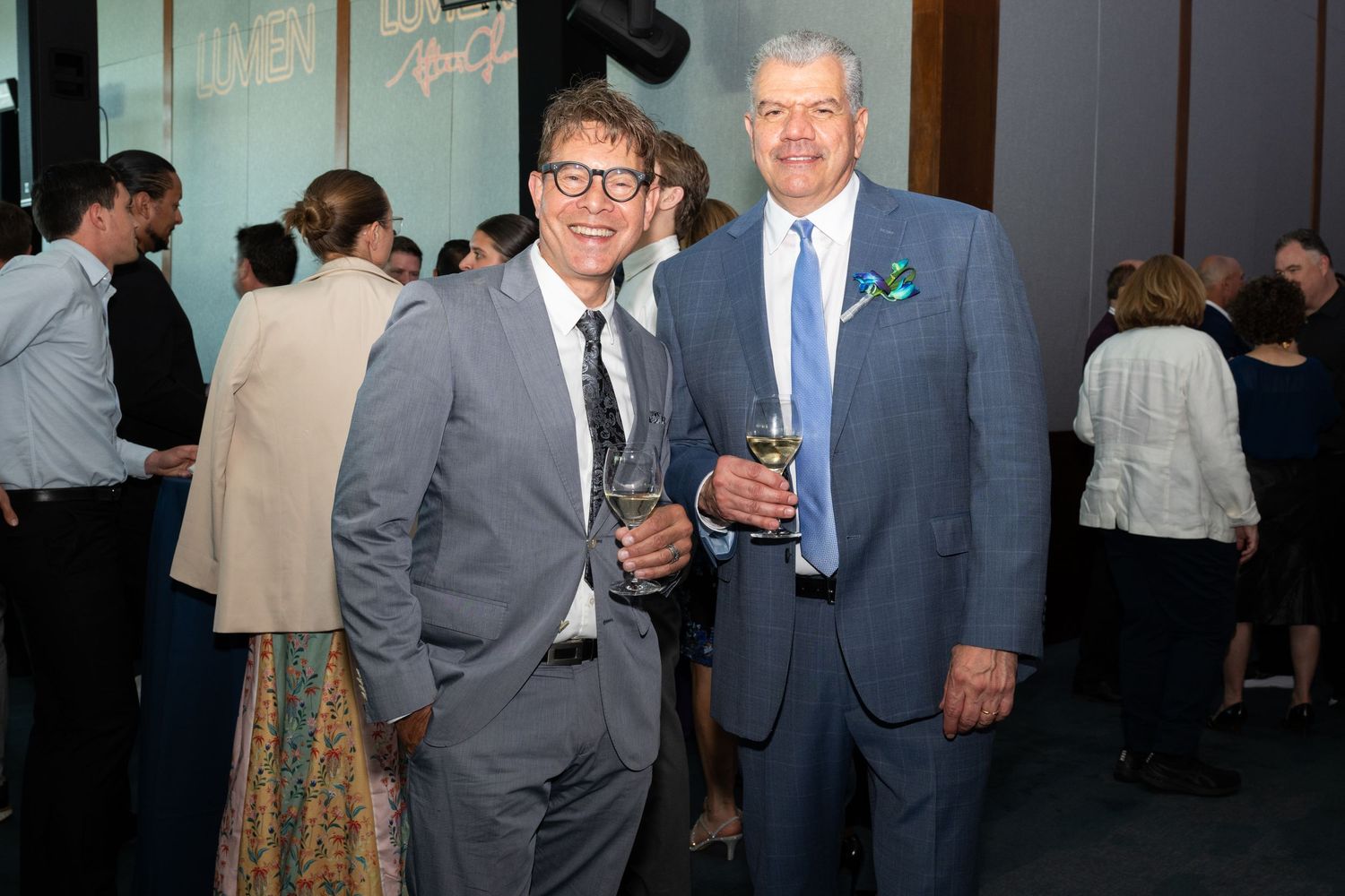 Two men in suits smile and hold drinks at an elegant evening reception with guests mingling in background.