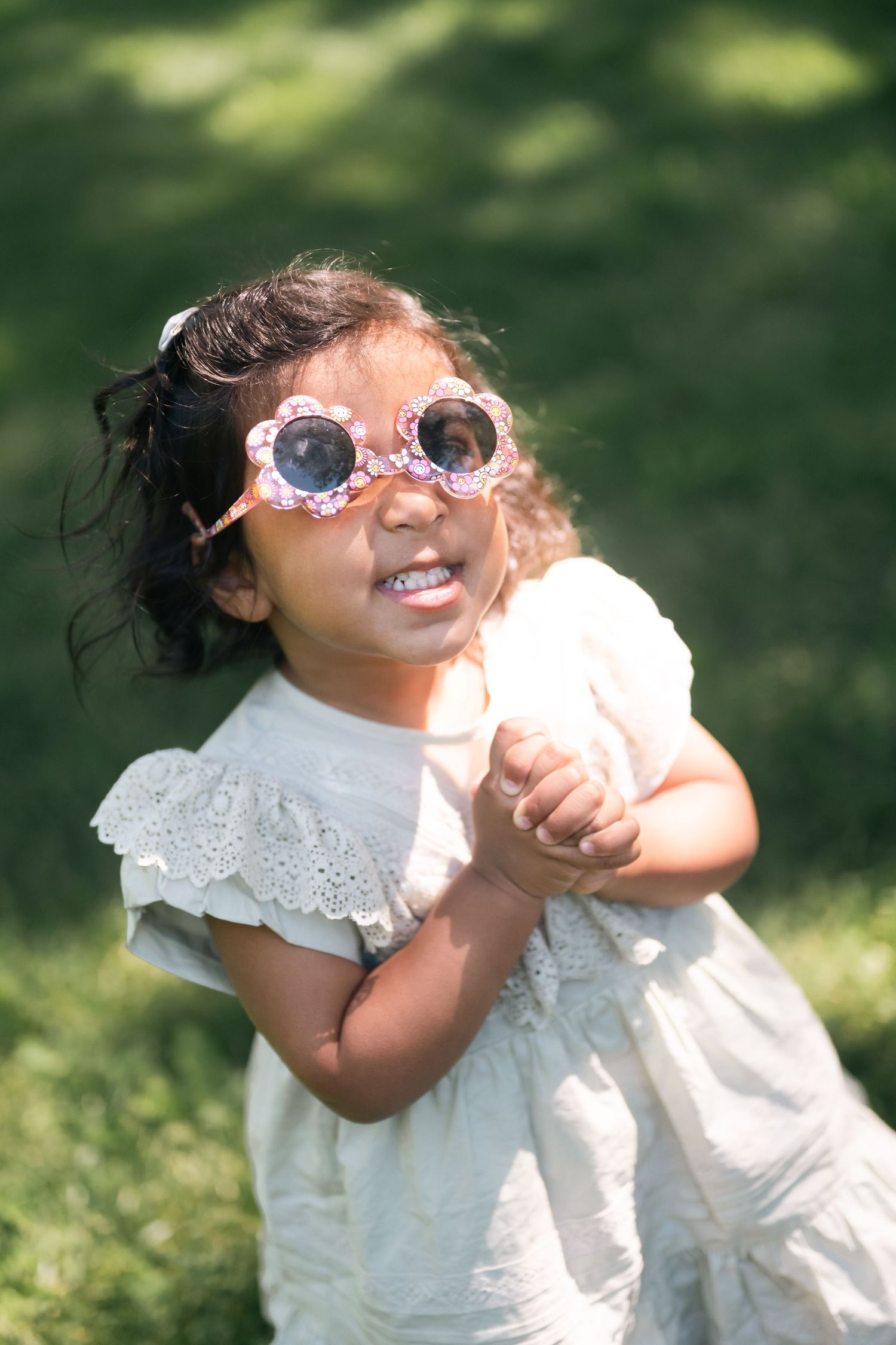 Cute toddler girl wearing floral sunglasses and white dress playing outdoors on green grass.
