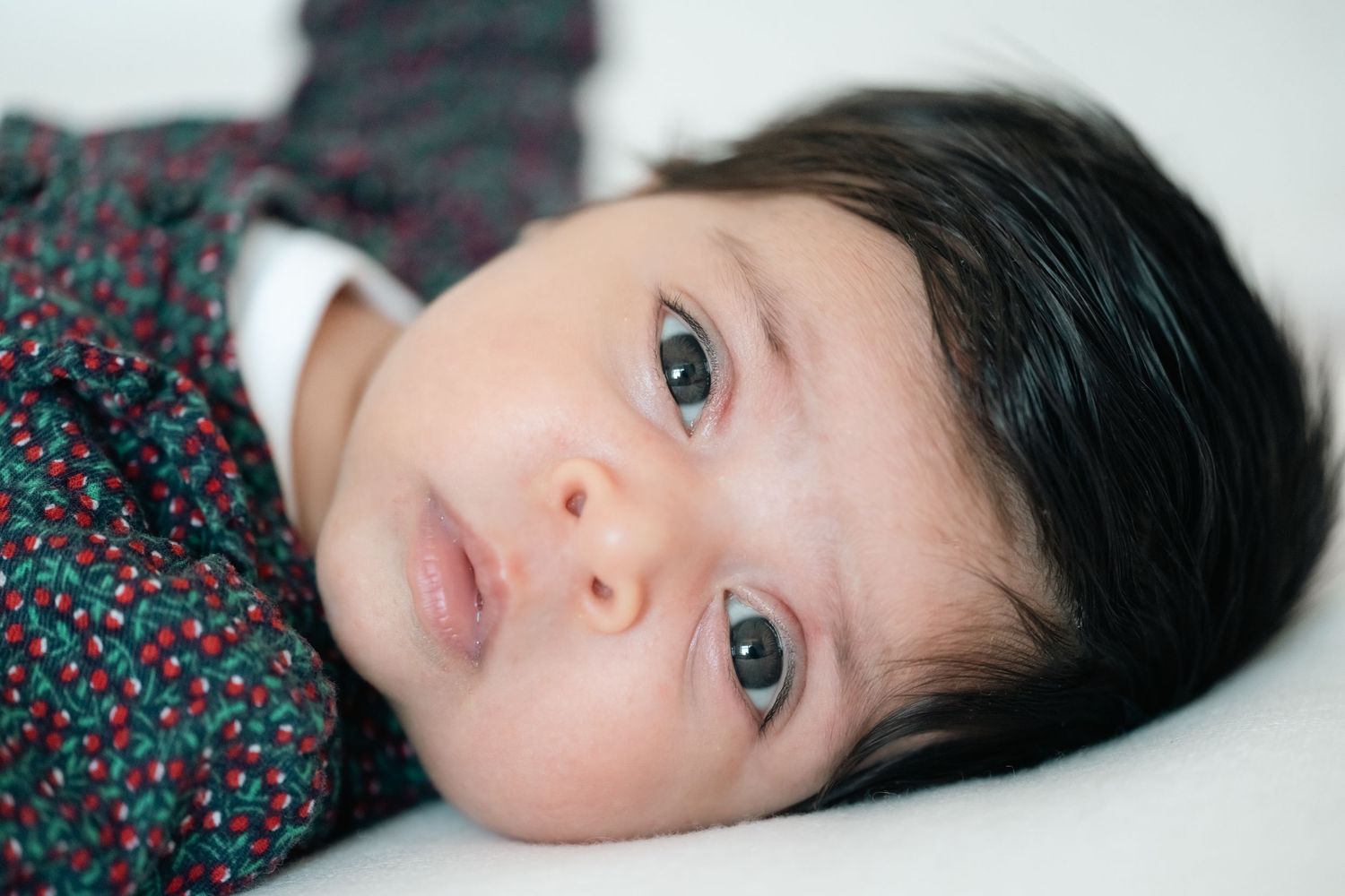 Close-up portrait of a newborn baby with dark hair and bright eyes lying on a white surface.