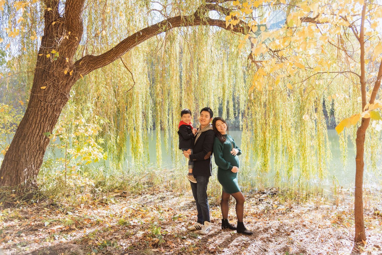 Family posing under a weeping willow tree in autumn park with colorful fall foliage near a lake.