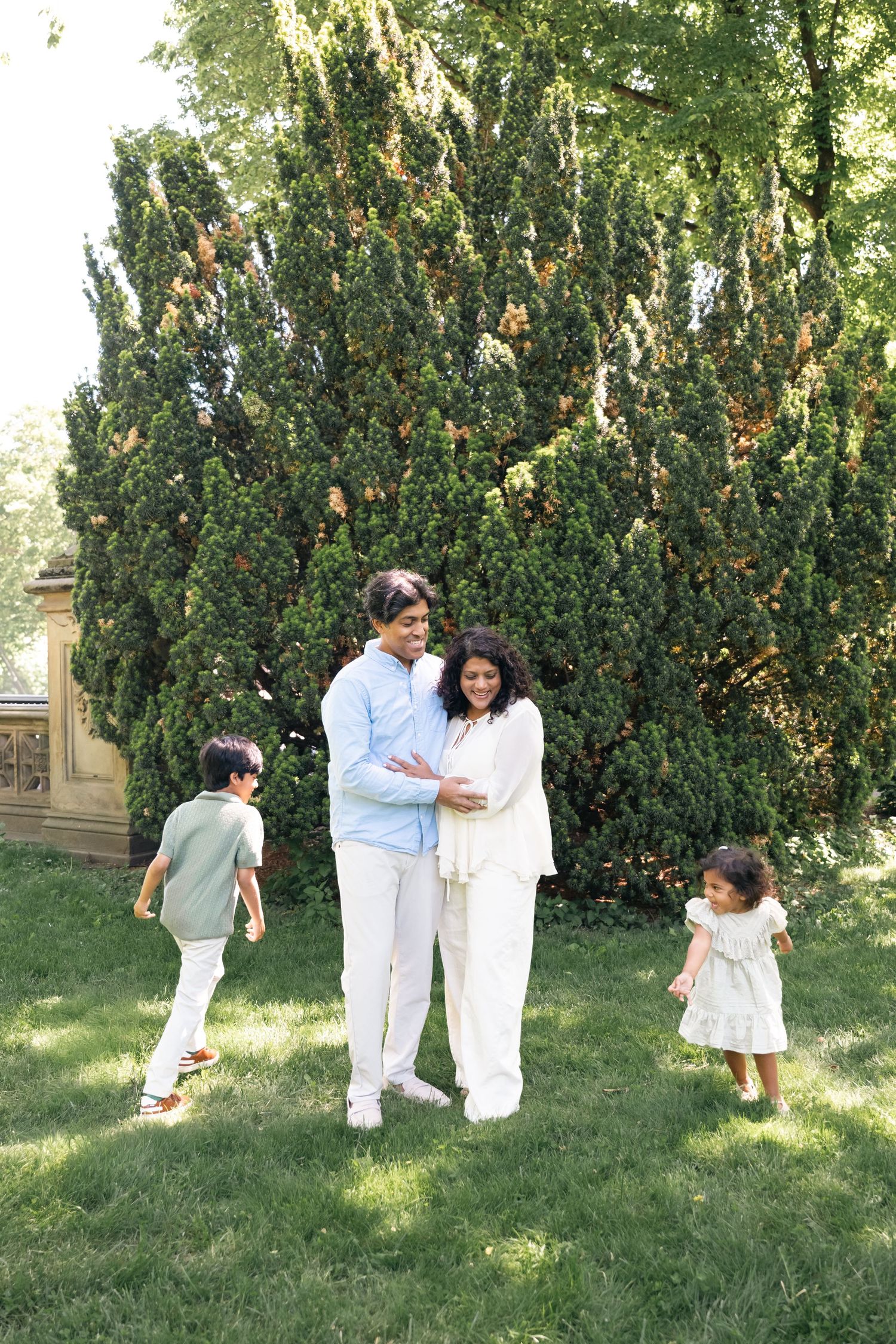 A happy family of four poses outdoors on green grass with a large tree backdrop on a sunny day.