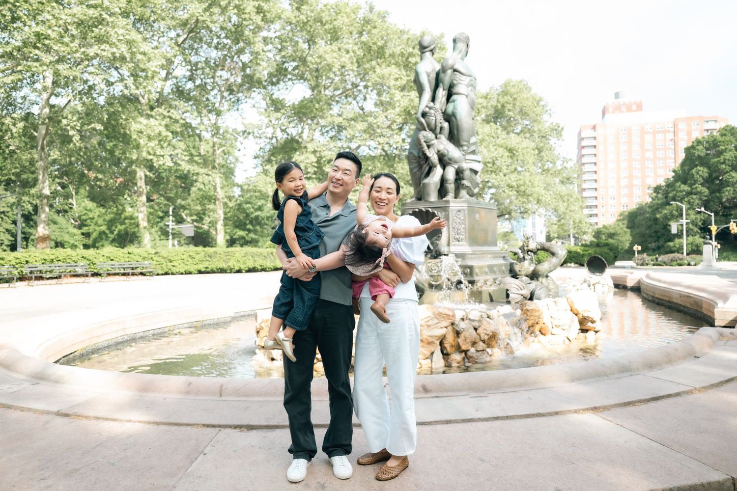 Family of four posing happily by a fountain in a park, with two children being held by their parents.