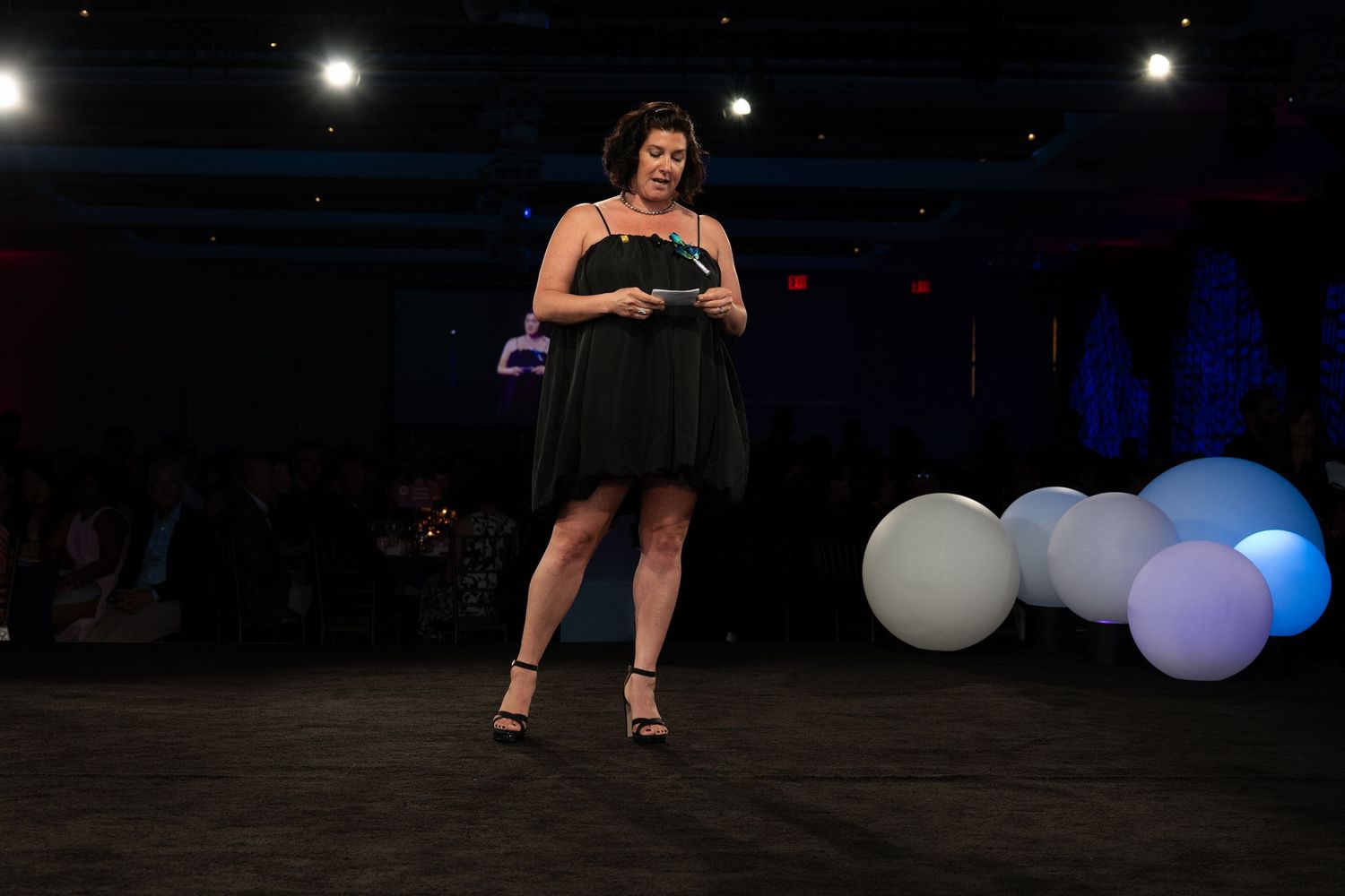 Woman in black dress presents on stage with decorative balloons at an indoor evening gala event.