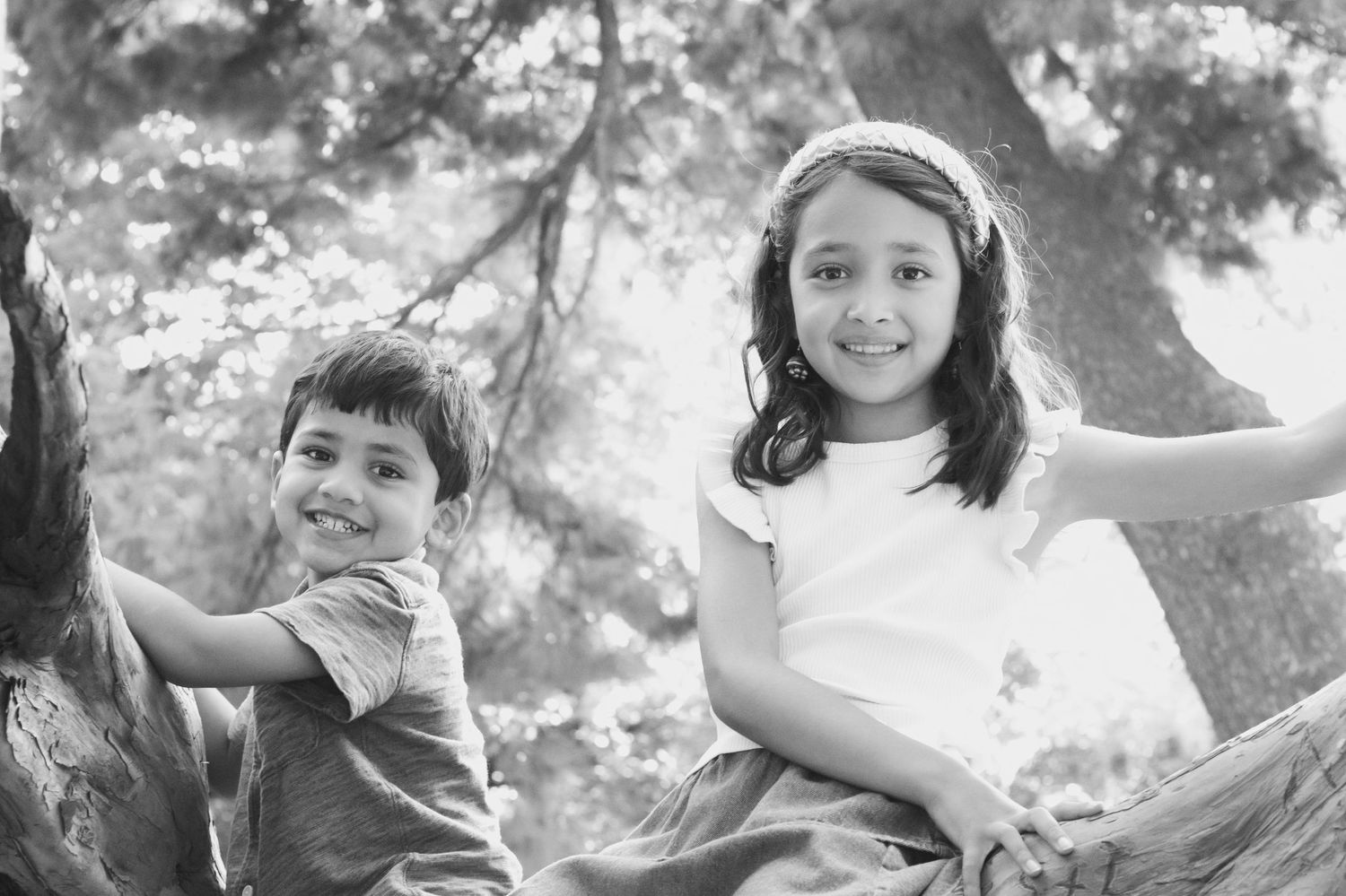 Two children sit together on a tree branch in a black and white outdoor portrait.