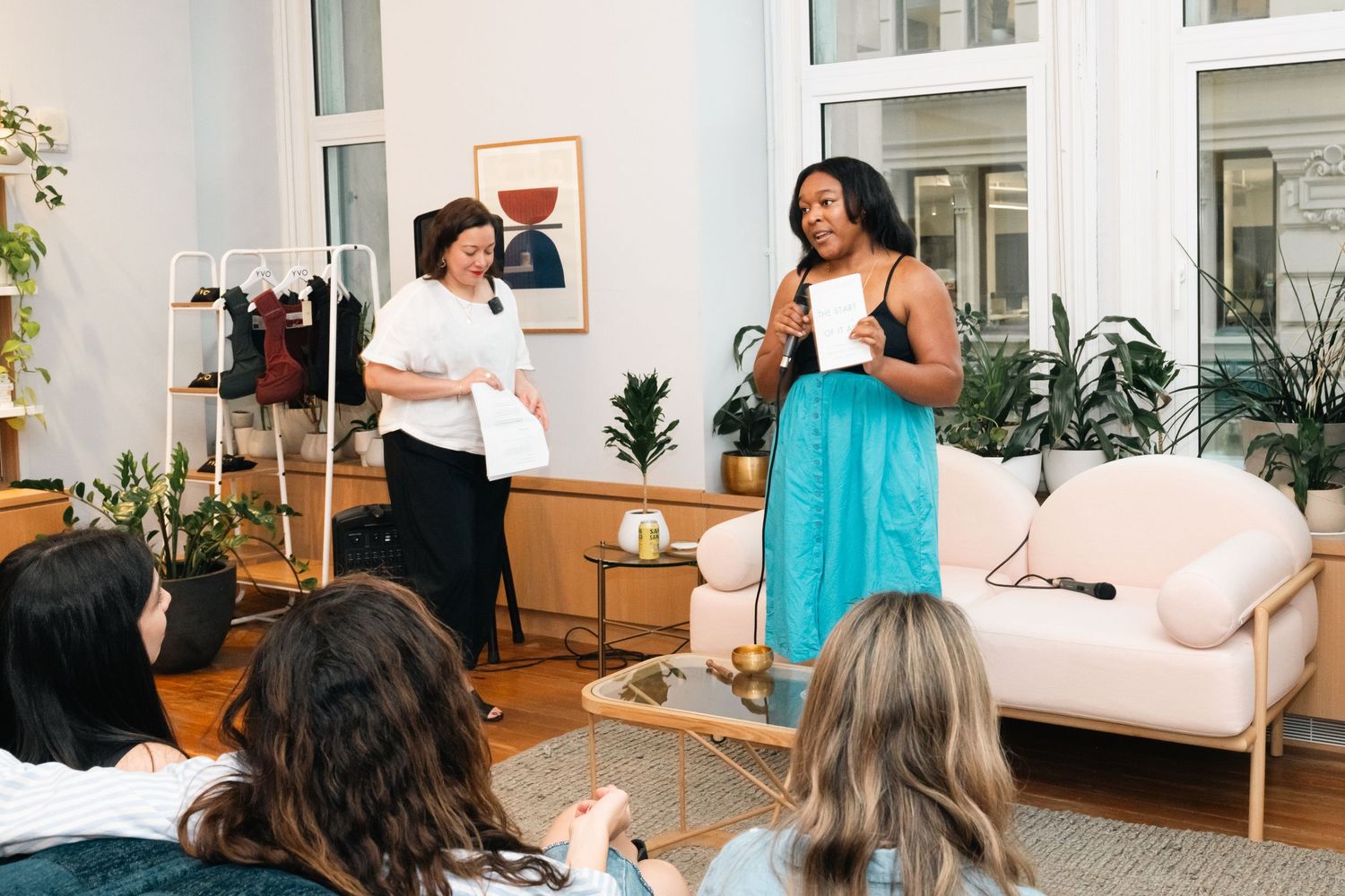 Two women presenting together in a bright modern living room space with plants and a pink sofa.