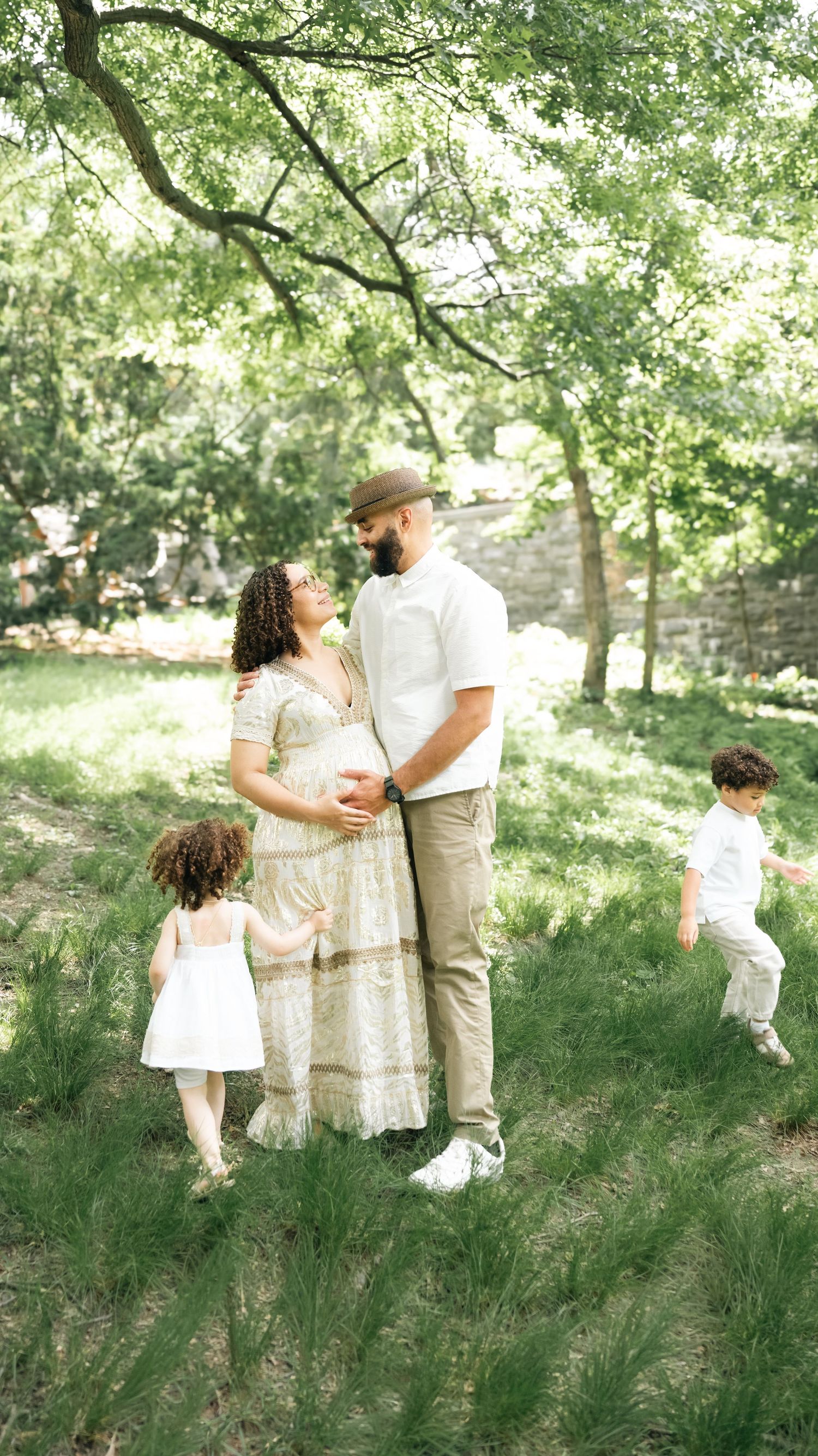 Pregnant woman in floral dress with husband and two children enjoying a sunny outdoor family photo session in a lush green park.