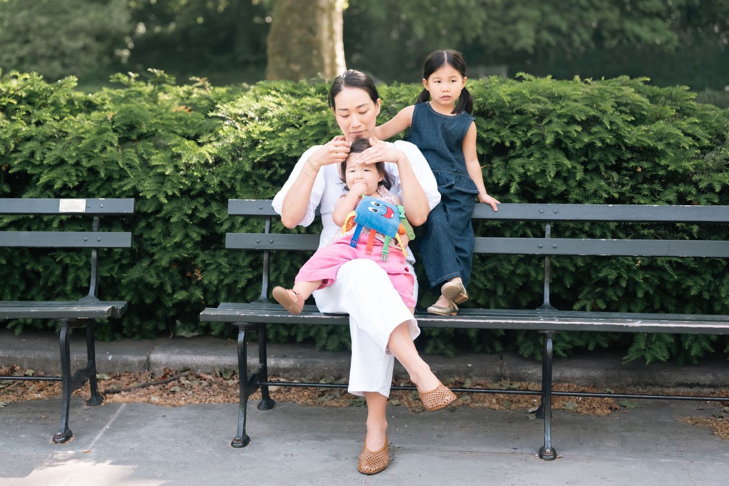 A woman sits on a park bench with two young girls climbing on her, surrounded by green hedges.