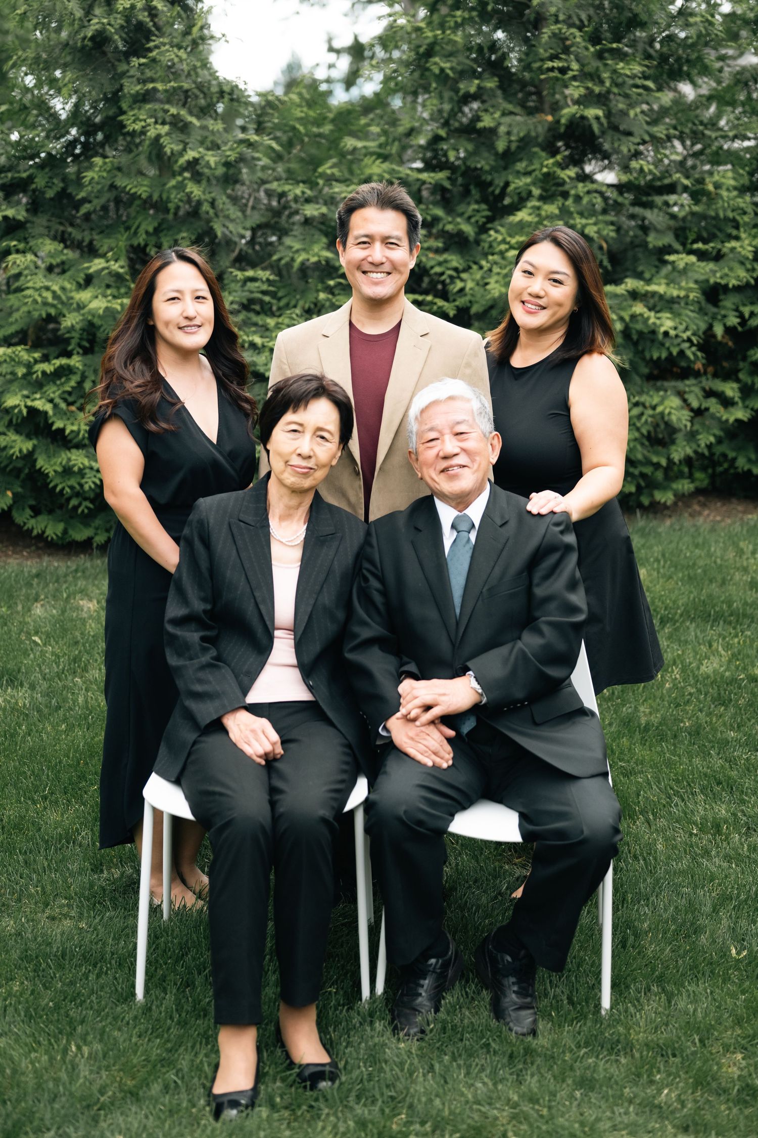 Multi-generational family members pose together on chairs in a garden setting wearing black formal attire.