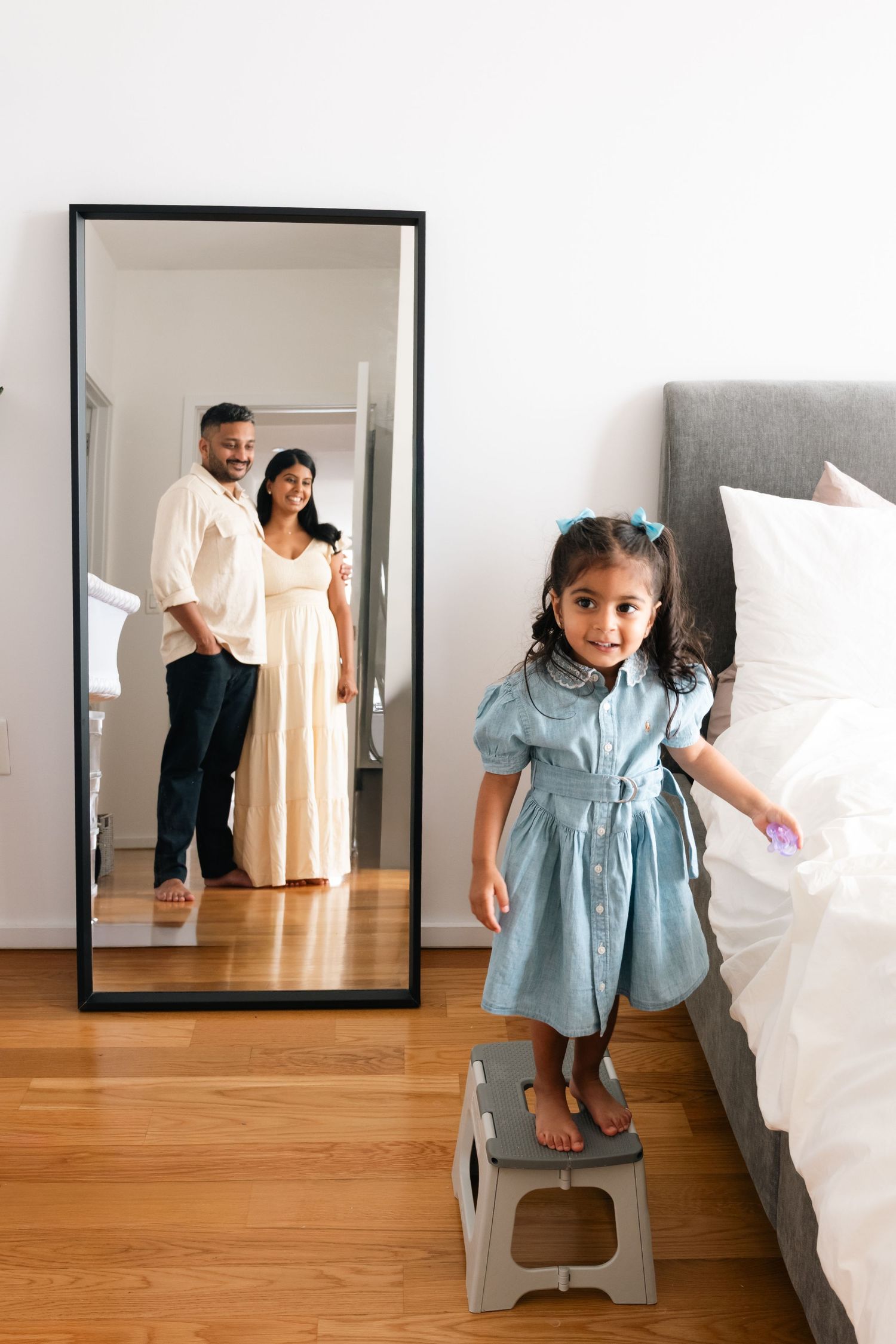 A daughter in a blue dress stands on a stool next to a white bed in a room with hardwood floors. Looking at her parents. Reflection shot