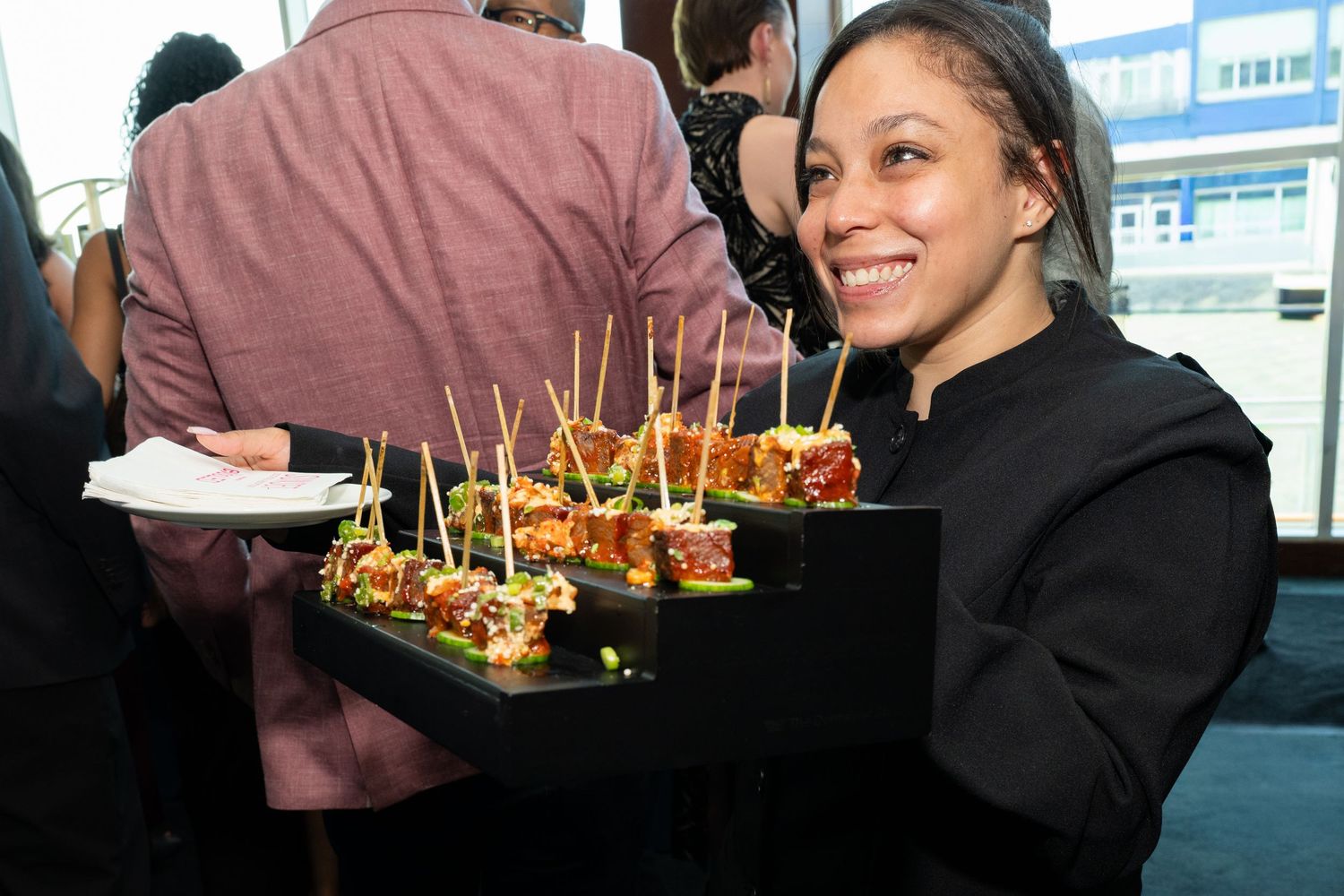 A smiling server in black attire holds a tray of elegant appetizers with decorative picks at a catered event.