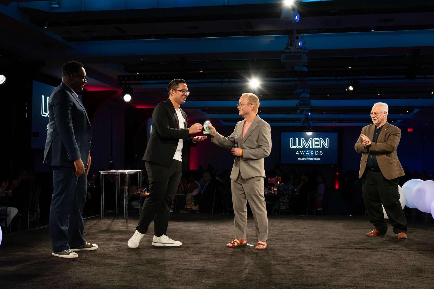 Four men on stage at the Lumen Awards ceremony, with two shaking hands during a formal presentation.