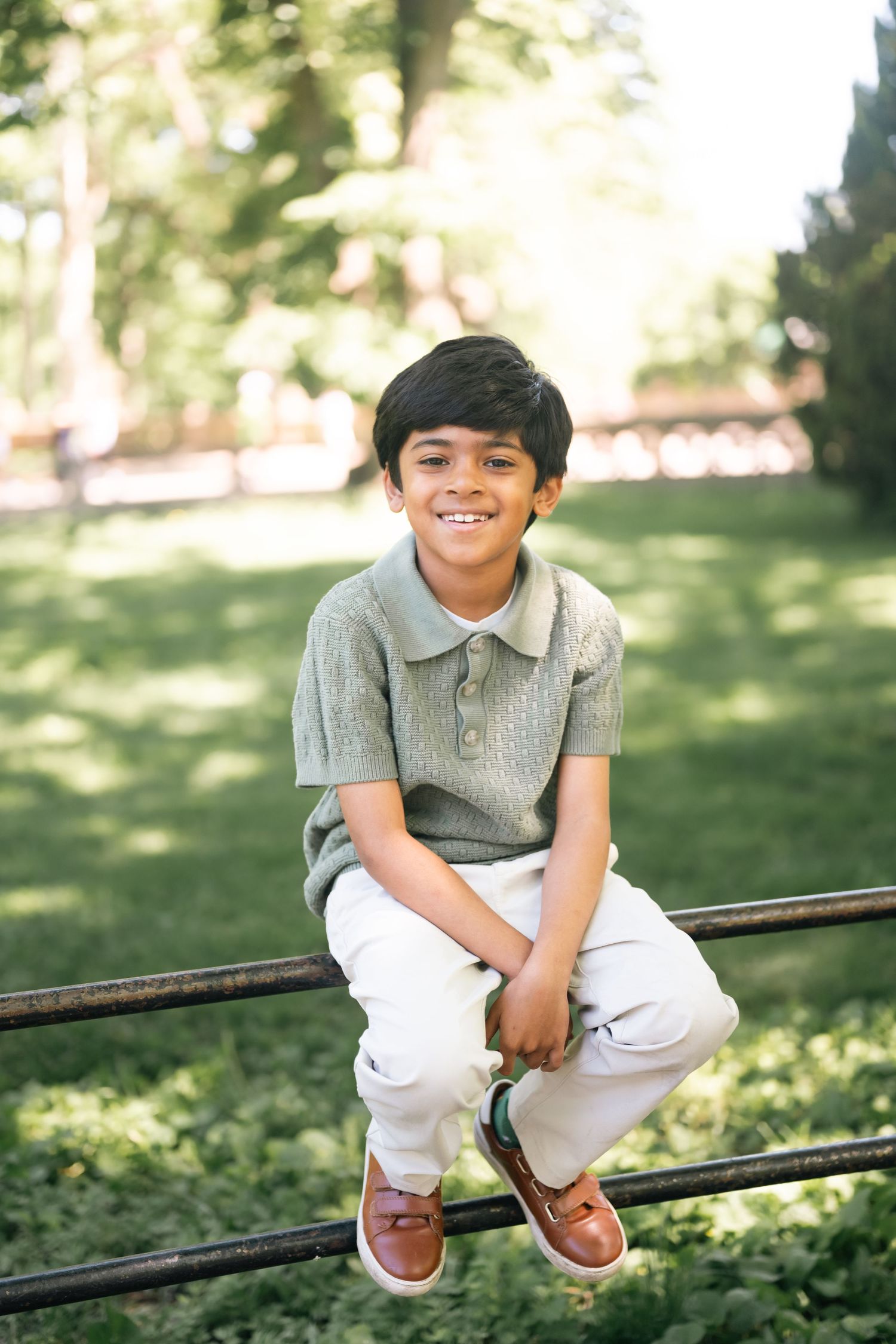 A smiling young boy sits on a metal fence in a sunny park, wearing a green polo shirt and white pants.