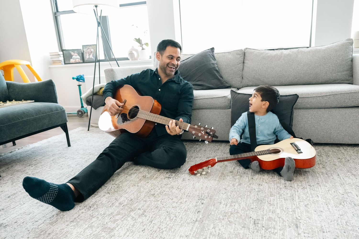 Father and child sitting on living room floor playing acoustic guitars together, smiling and bonding over music.