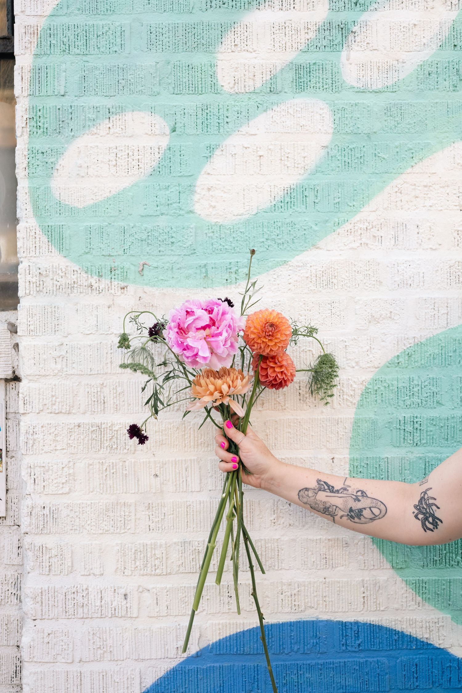 Hands holding colorful flower bouquets against a turquoise and white painted brick wall.