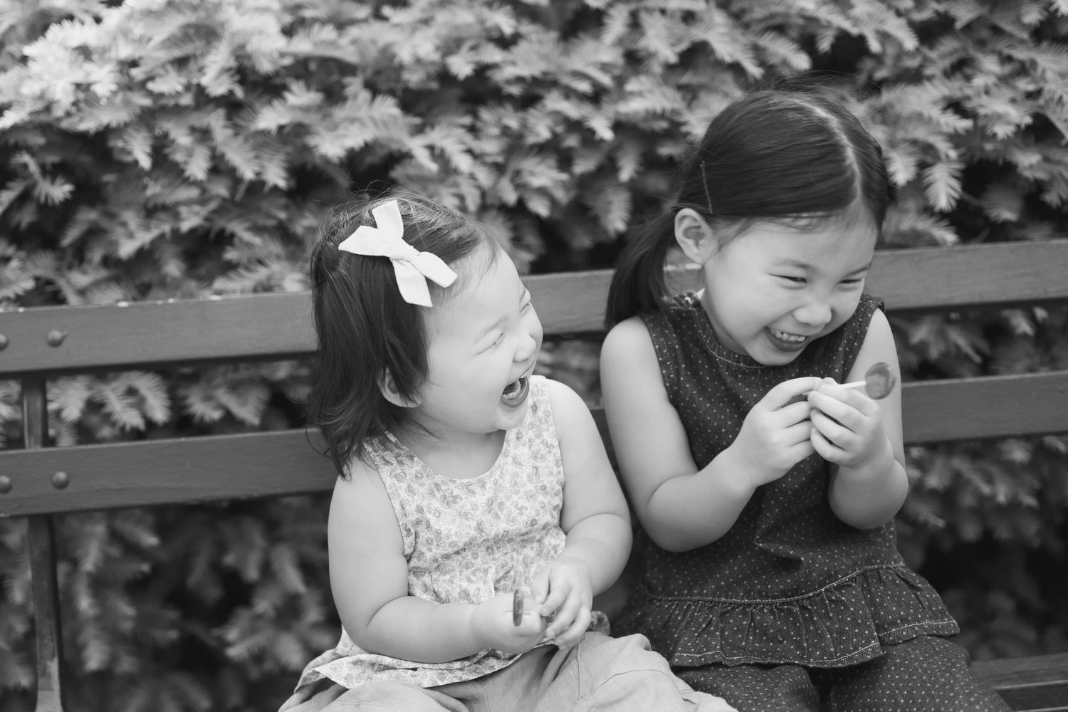 Two young Asian girls laughing joyfully together on a park bench in black and white photo.