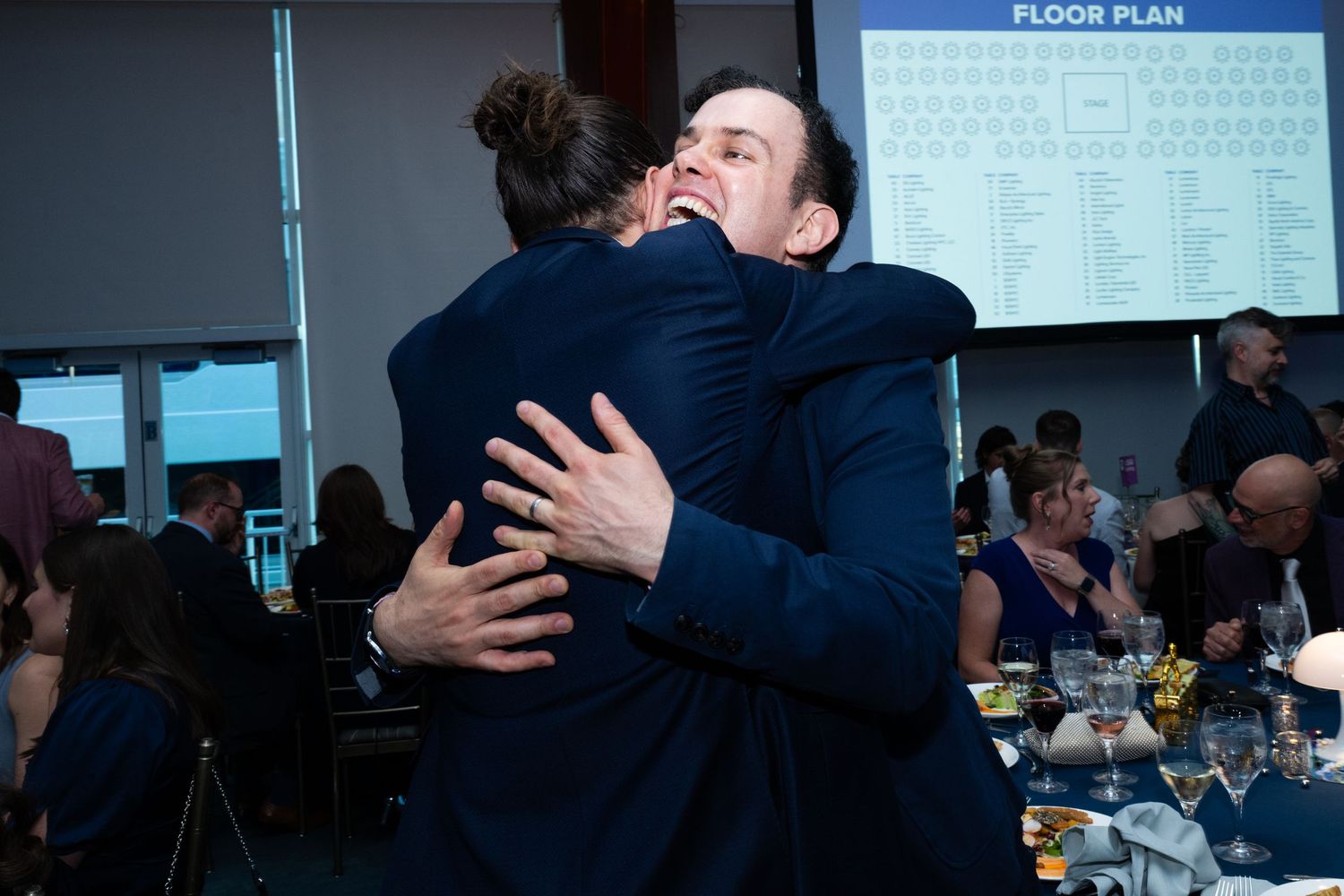 Two guests share a warm embrace at a formal banquet dinner event with seating chart visible.