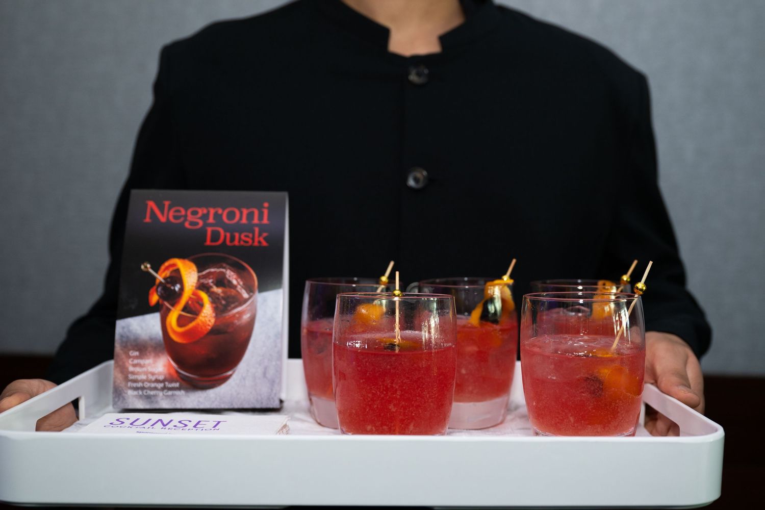 Server holding a tray with Negroni Dusk cocktails and cookbook at an elegant event.