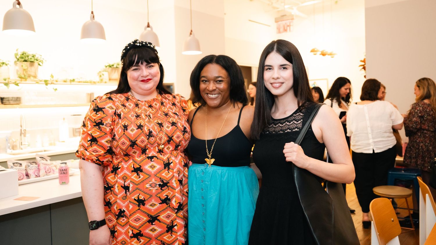 Three women smiling together at a beauty salon event, wearing colorful outfits in a bright modern interior.