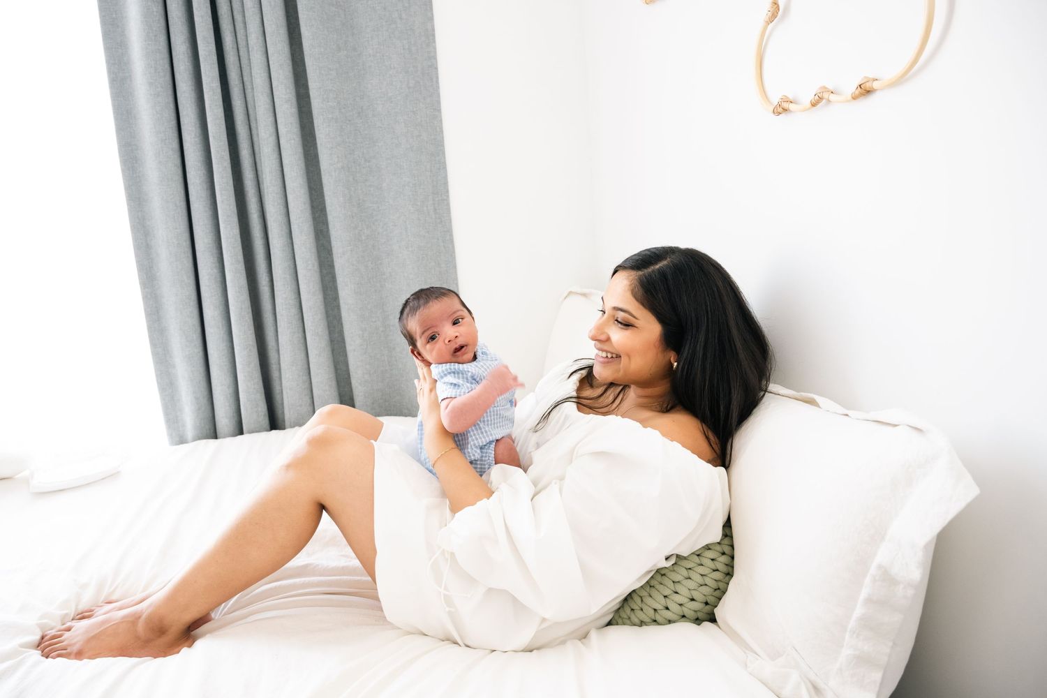 Mother and newborn relaxing together on white bed with grey curtains.