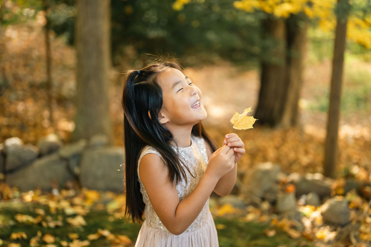 Child in white dress holding yellow flower in autumn forest setting.