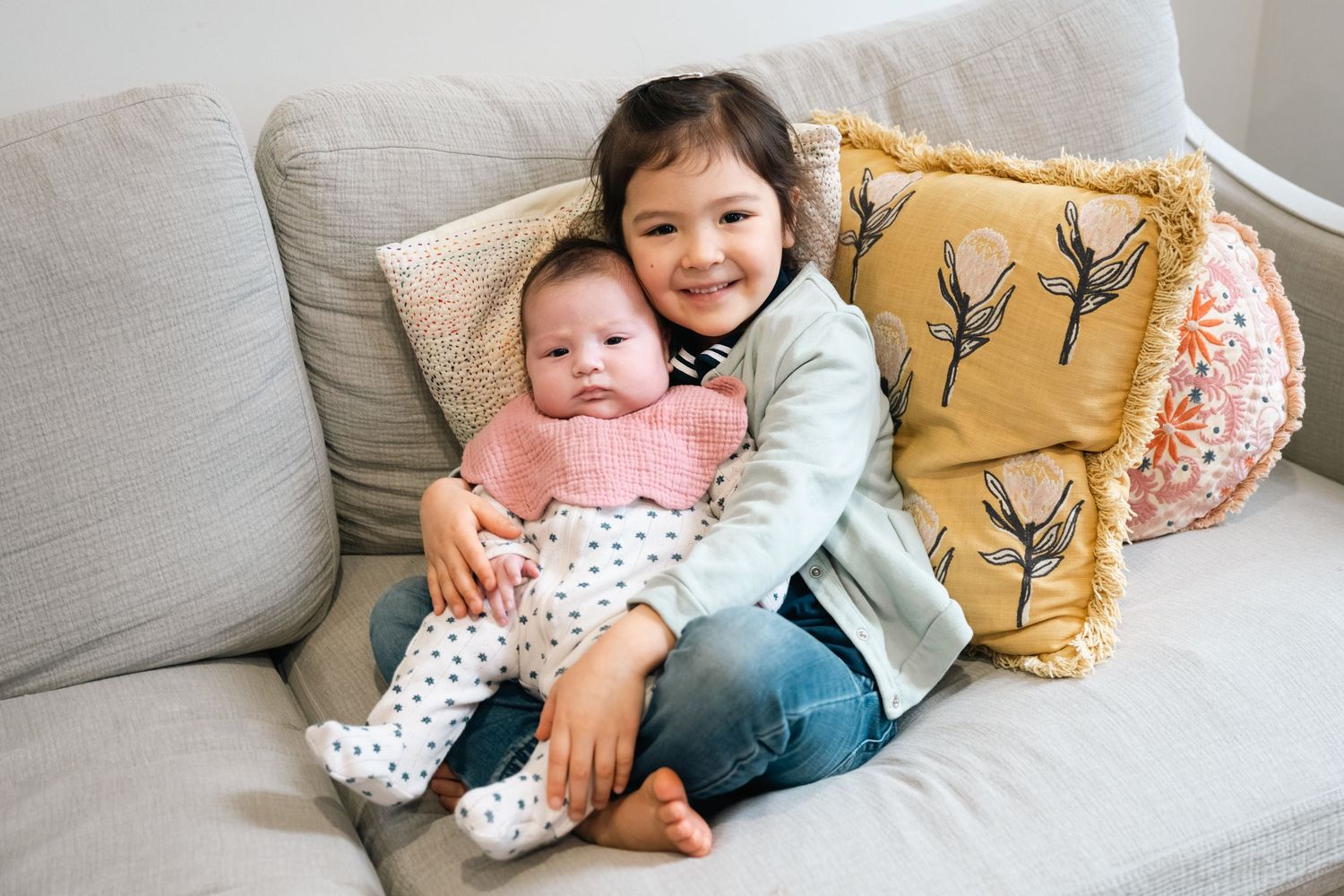 Two children share a sweet moment while sitting on a grey couch with yellow and floral accent pillows.