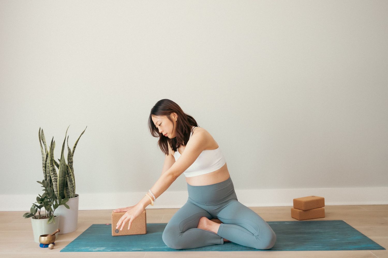 Woman practicing yoga stretches on a teal mat with wooden blocks and plants in a minimalist setting.