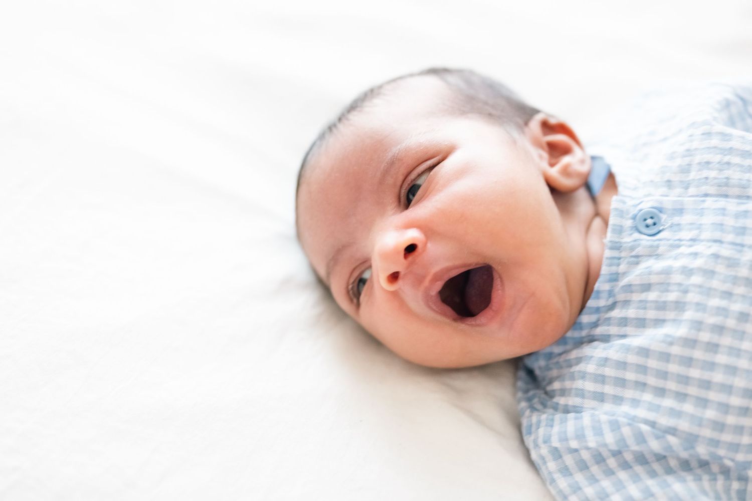 Adorable baby in blue checkered outfit yawning while lying on white bedding.