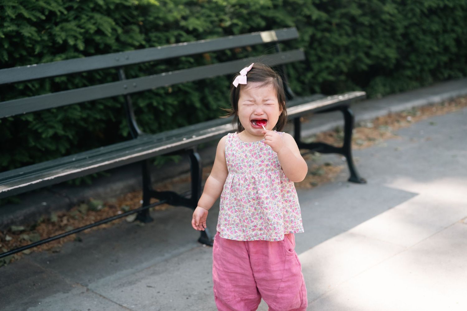A young toddler girl in a pink floral top crying outdoors near a park bench surrounded by green hedges.