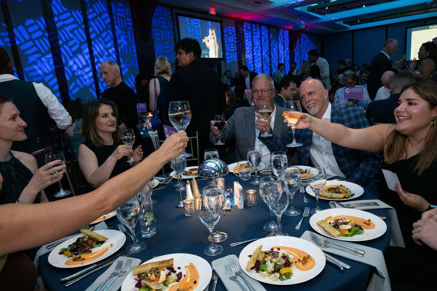 Guests raise wine glasses in a toast over plated dinner at an elegant blue-lit gala event.