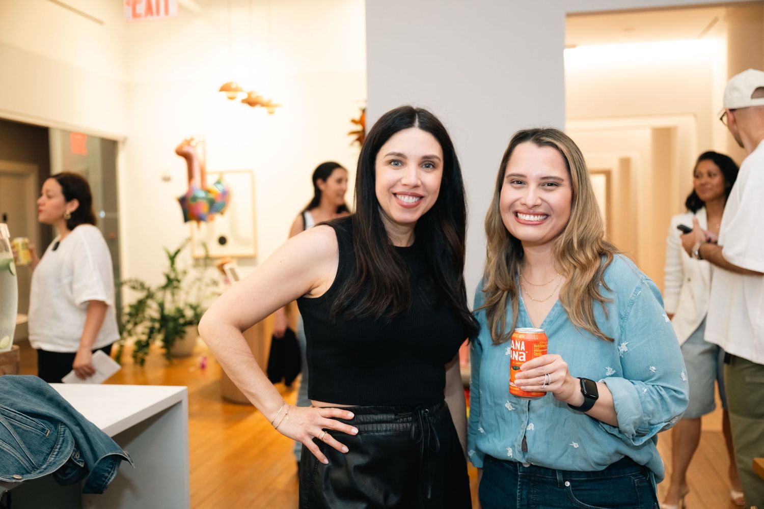 Two women smiling at an indoor social gathering event, one in black outfit, other in blue shirt holding orange drink.