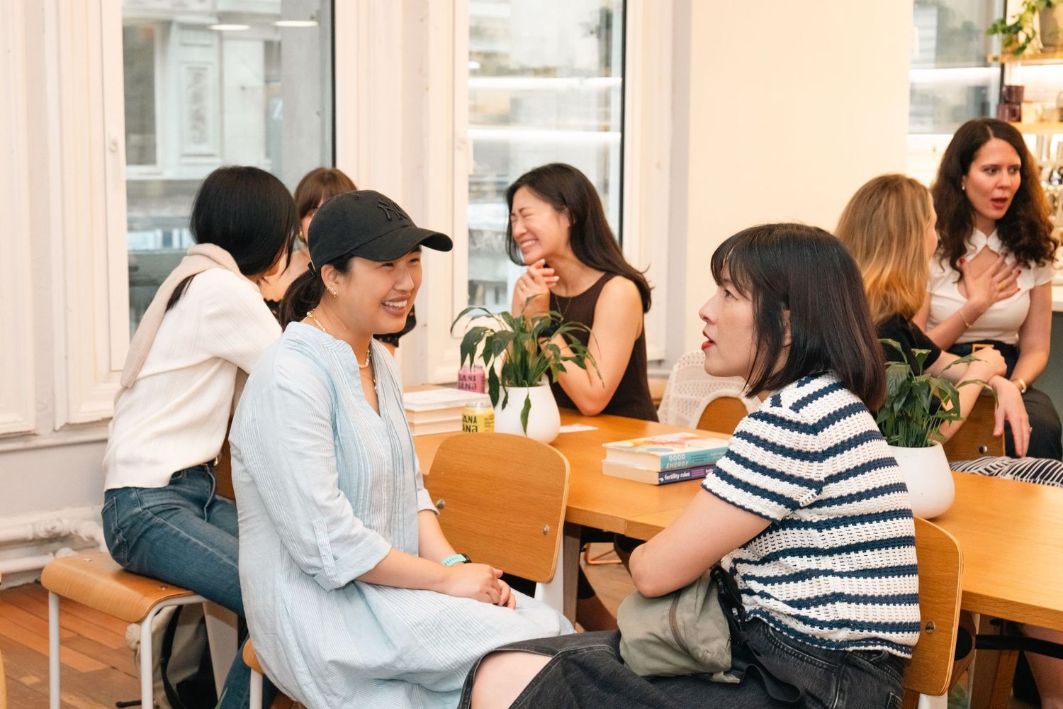 Diverse group of women laughing and talking together at wooden table in bright modern office space.