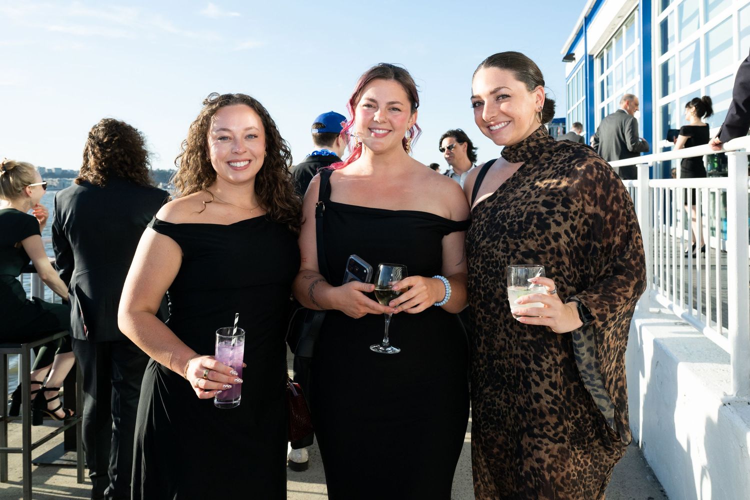Three women in elegant attire smile and hold drinks at an outdoor rooftop social event.