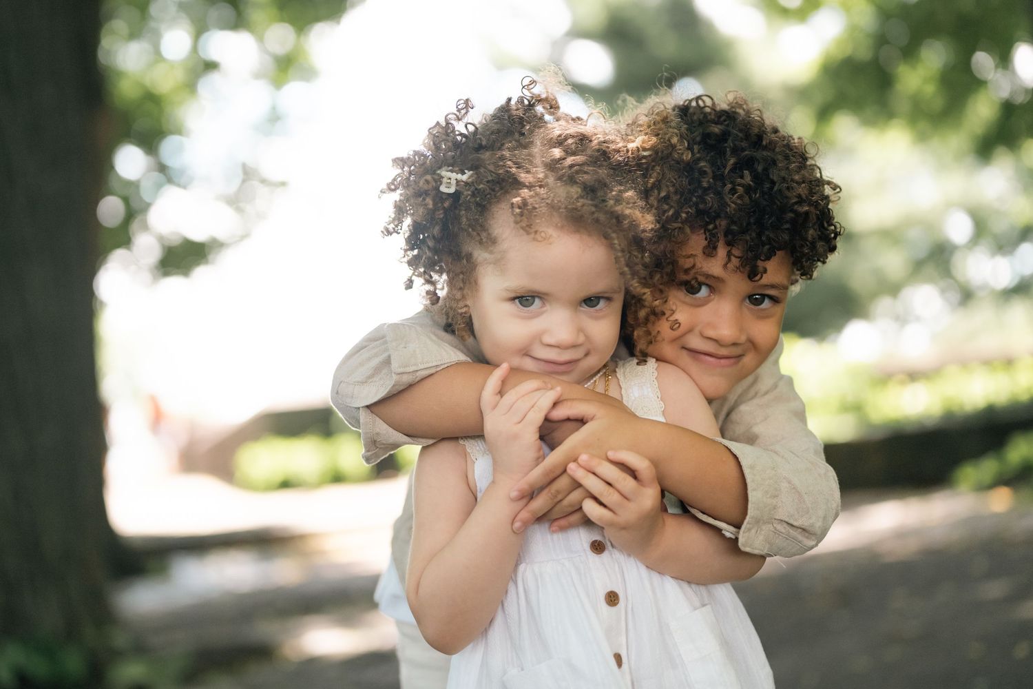 Two curly-haired toddlers hugging outdoors in a park, smiling joyfully in a heartwarming embrace.