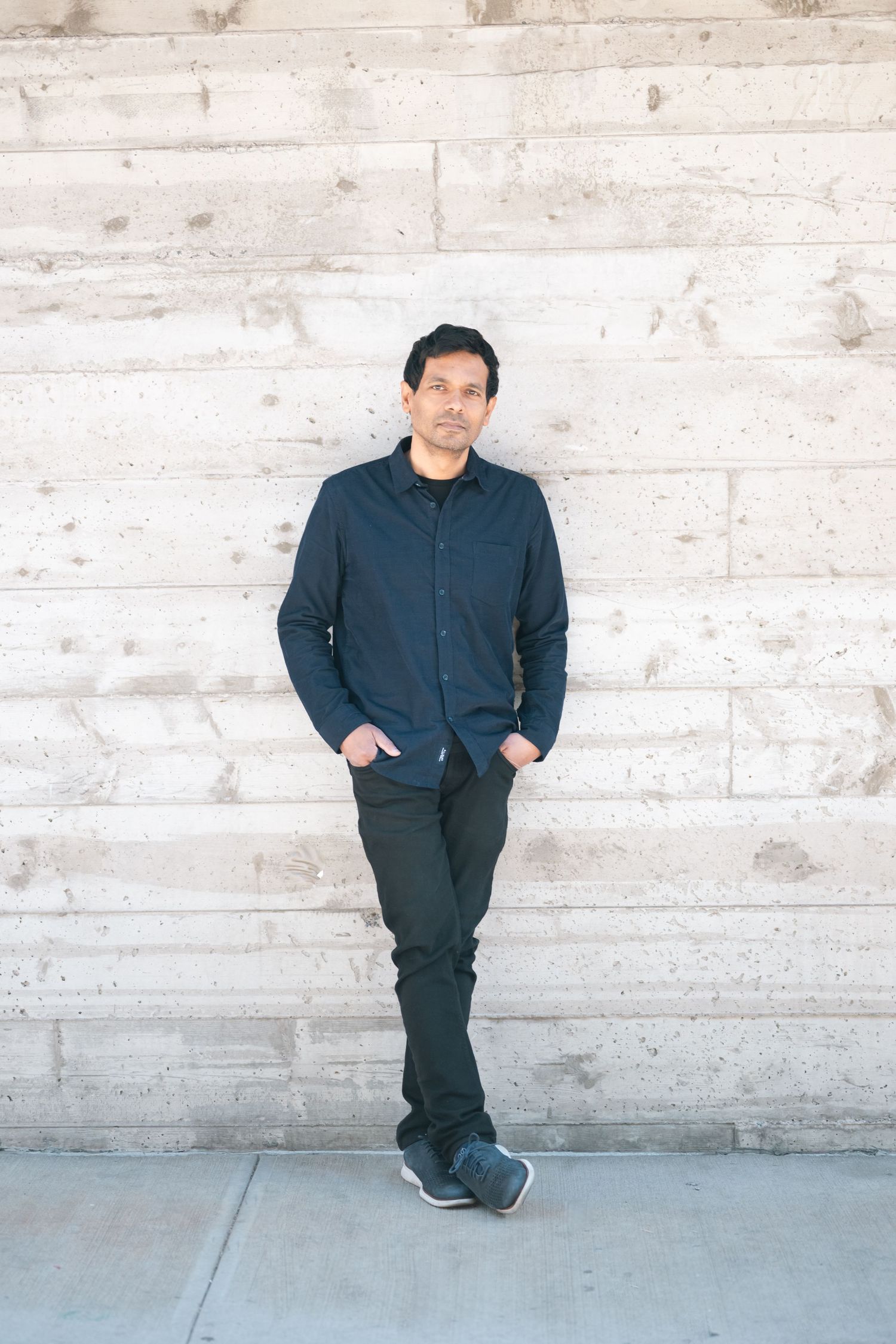 A man in a dark blue shirt and black pants leans against a concrete wall with hands in pockets.