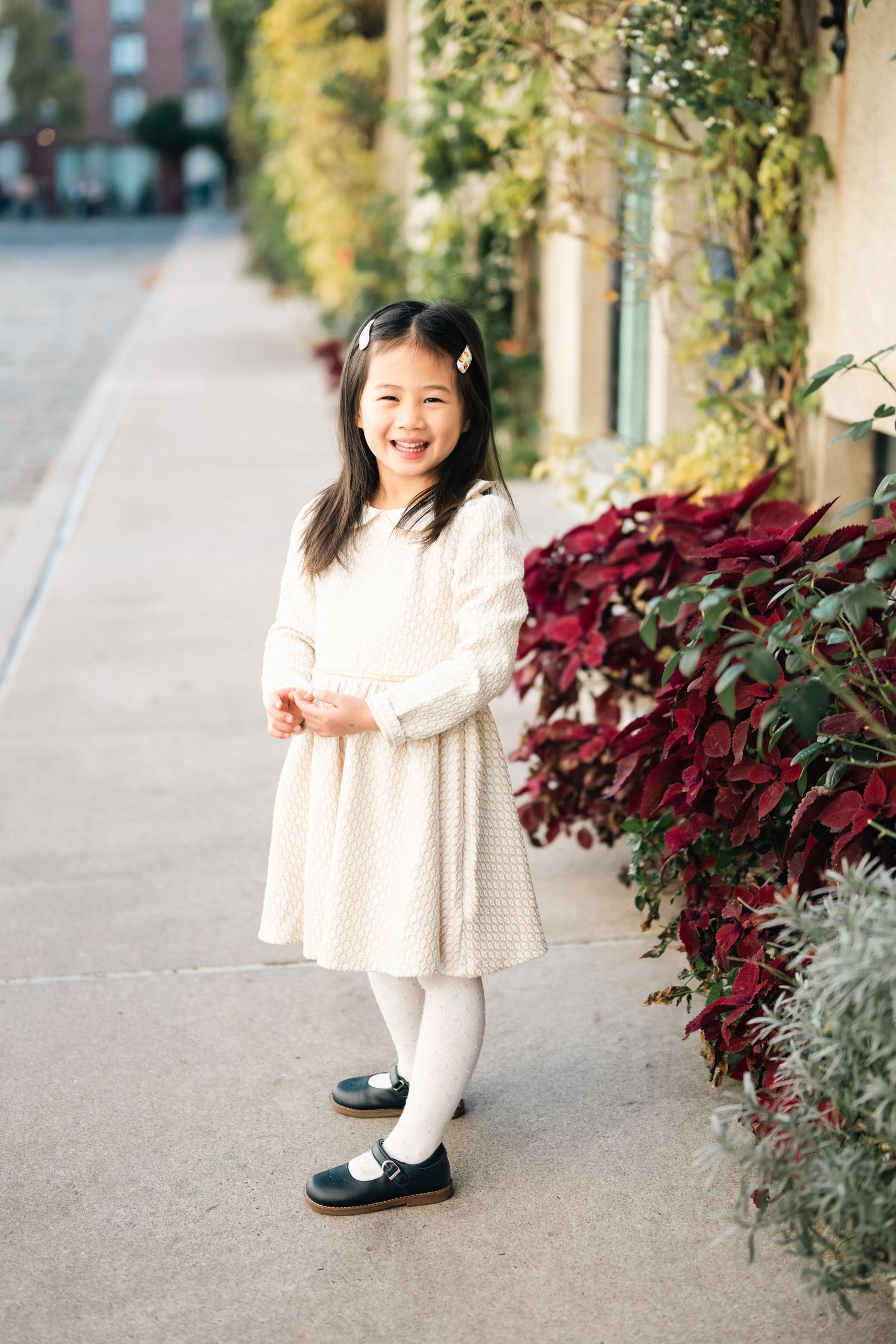 A child wearing a white dress poses by red and green plants along a walkway.