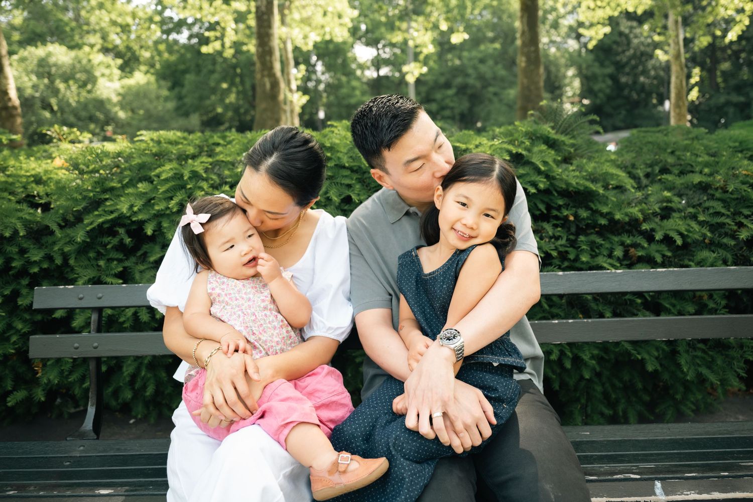 A happy Asian family of four sits on a park bench, parents kissing their two young daughters outdoors.