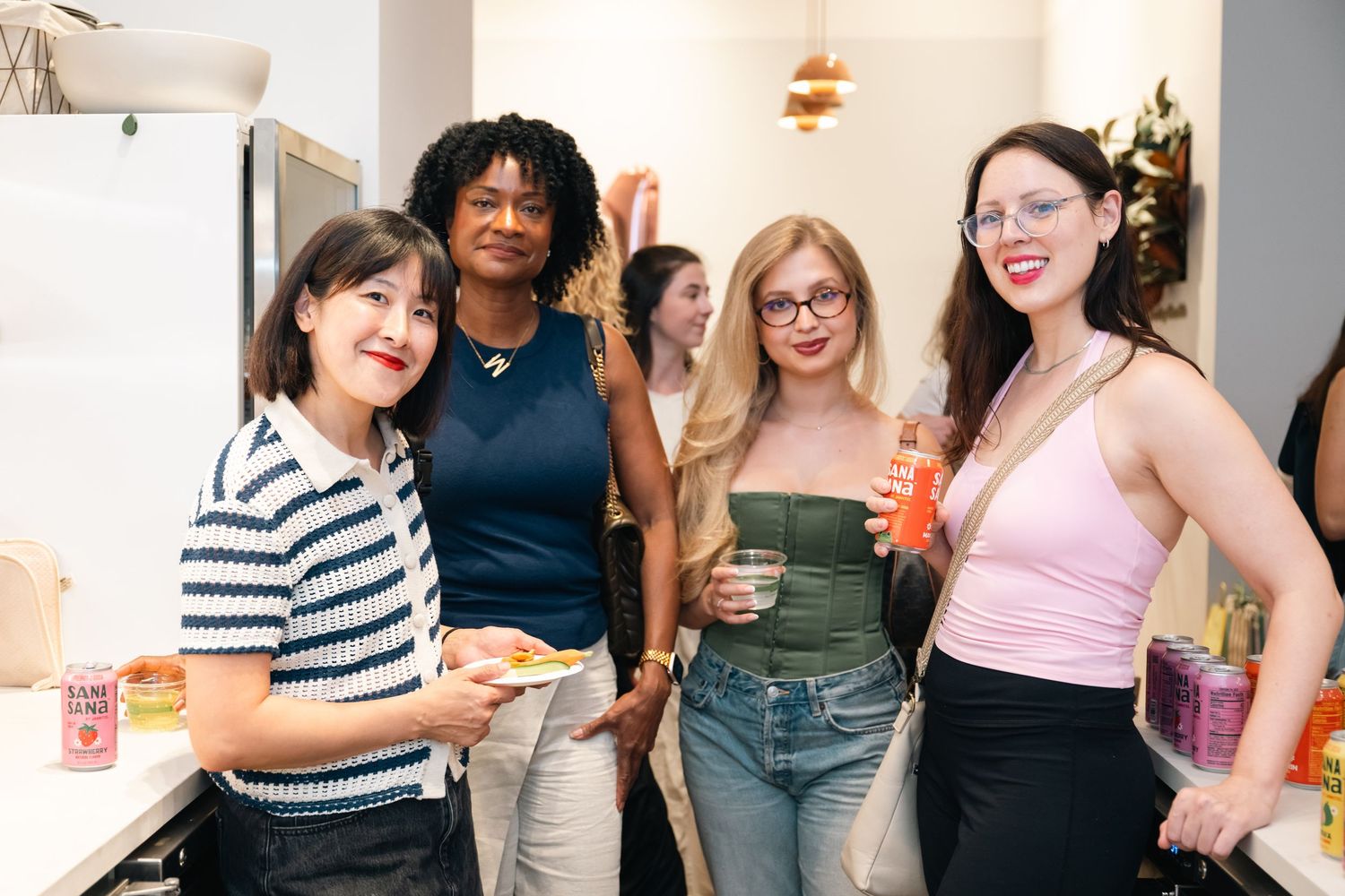 Four women smiling and socializing at an indoor gathering, holding drinks in a bright modern space.