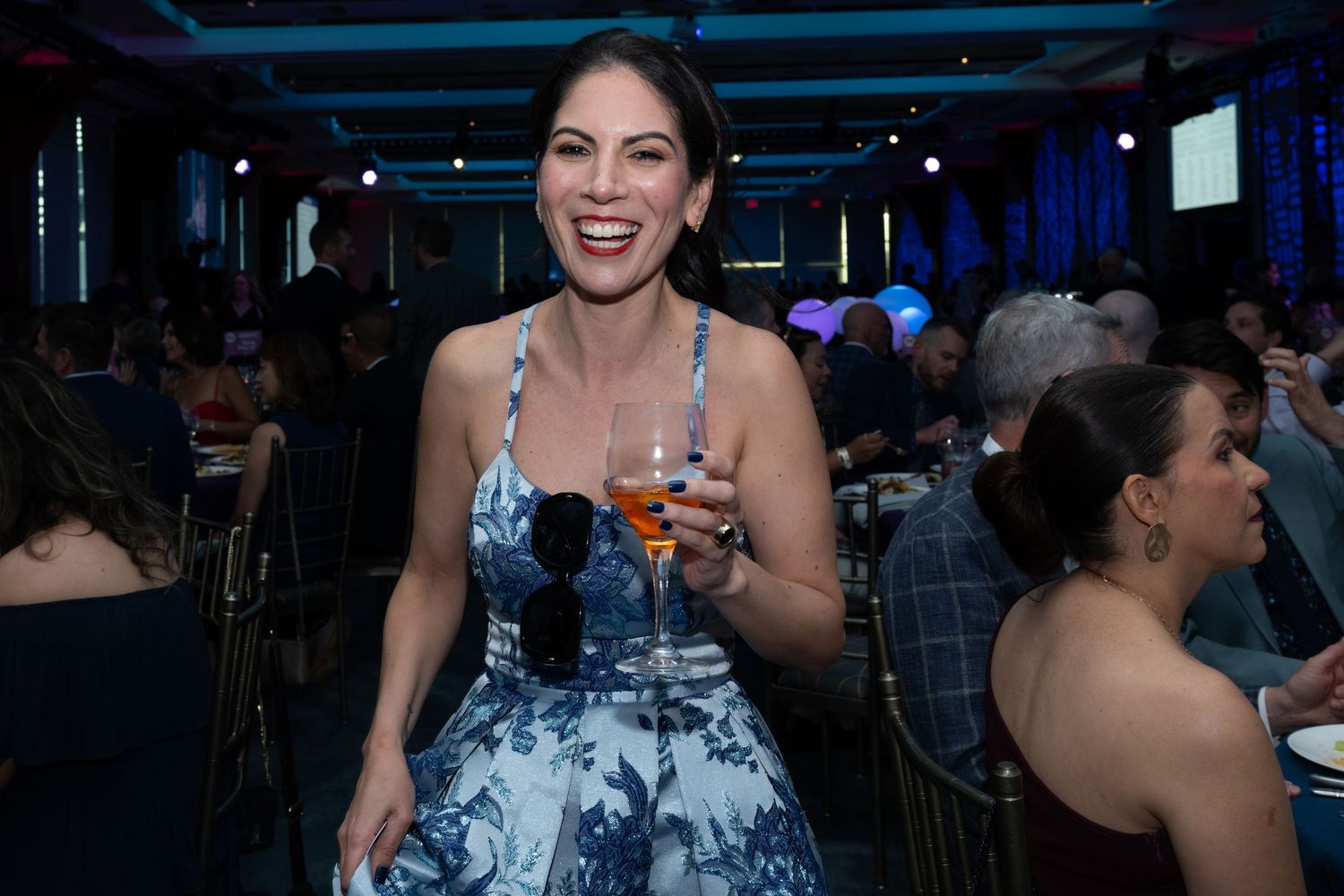 Smiling woman in floral dress holding a drink at a blue-lit gala dinner event.