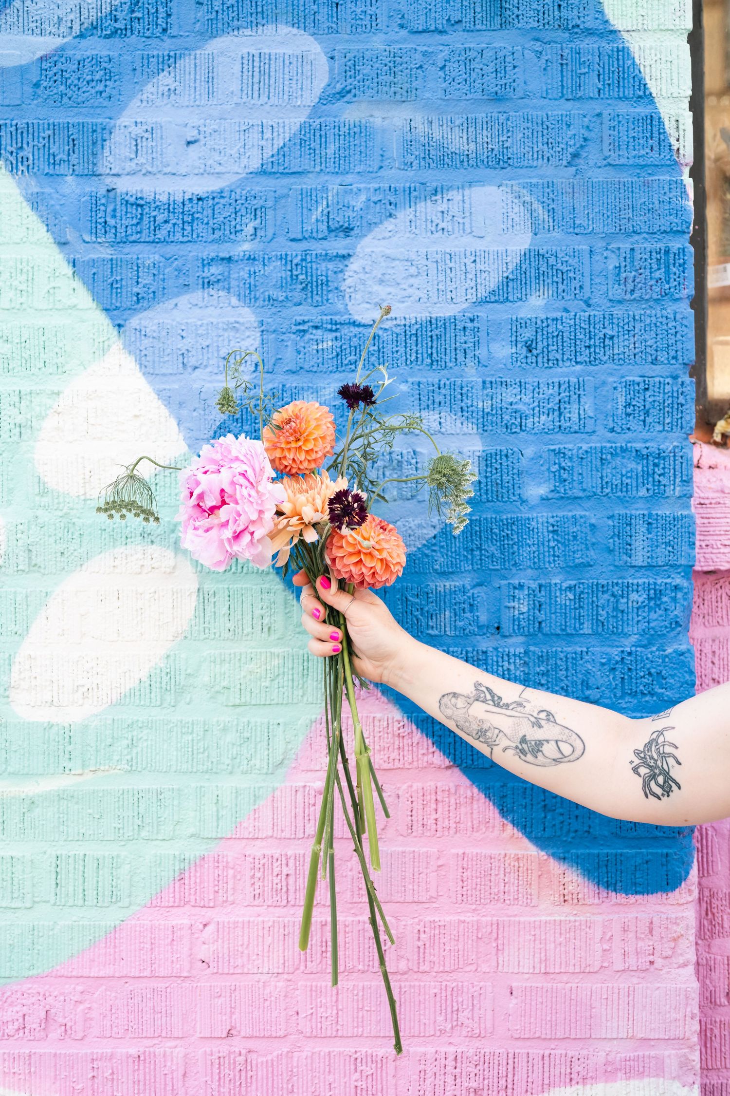 Hand holding pink and orange flowers against a blue and pink geometric mural.