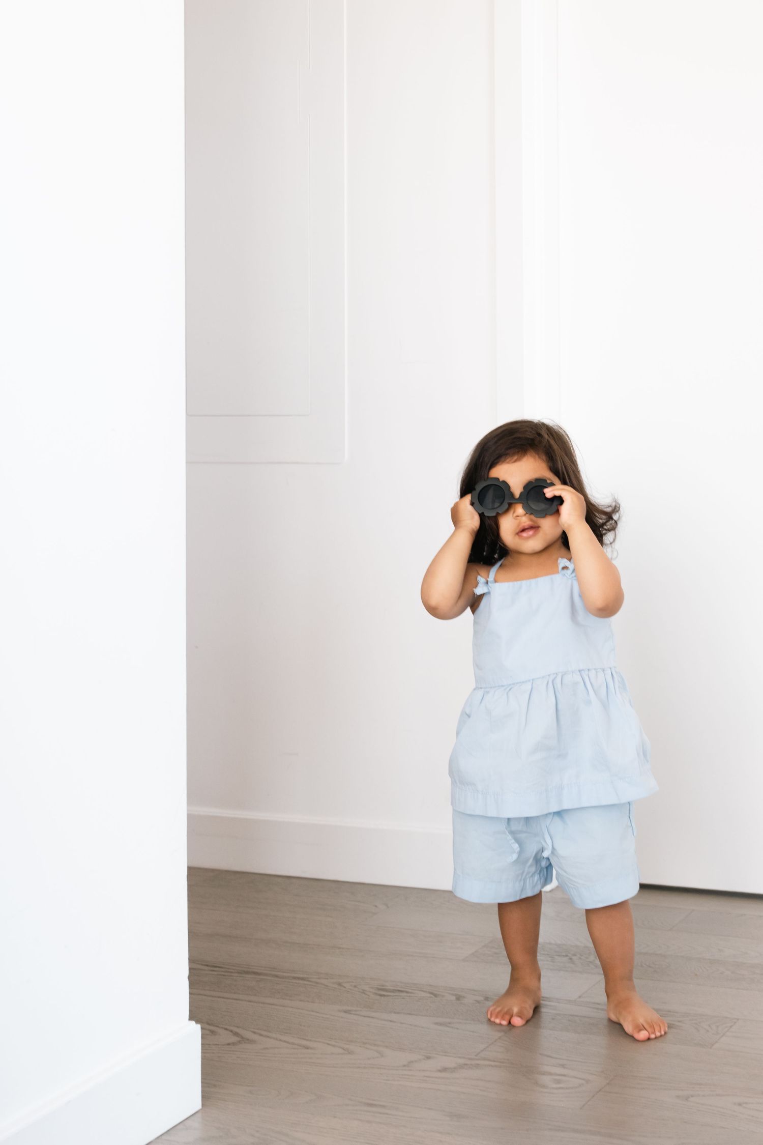 Young child in light blue outfit playing peek-a-boo against white walls.